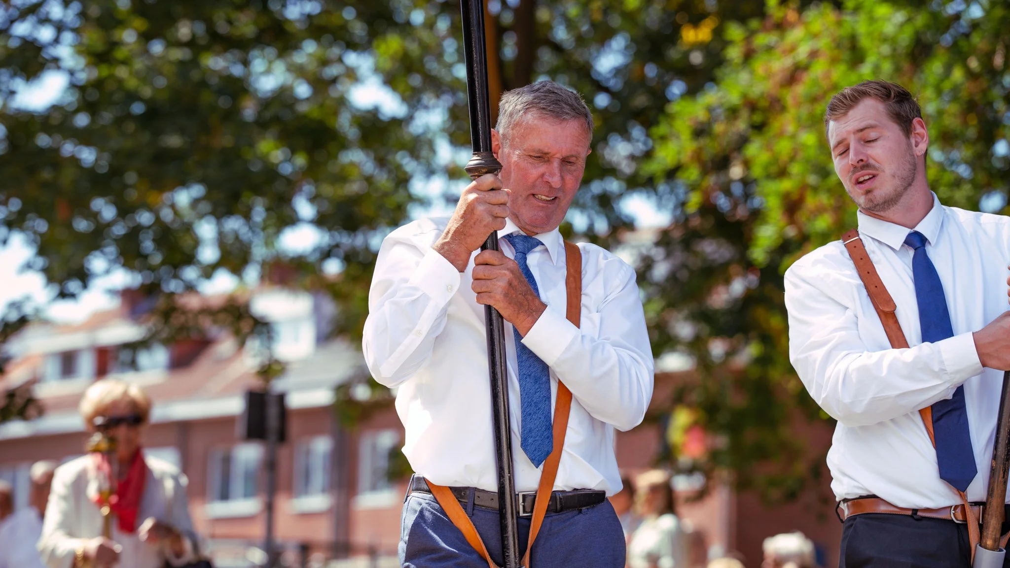 Two men in white shirts and blue ties holding black umbrellas, standing outdoors under green trees with a brick building in the background.