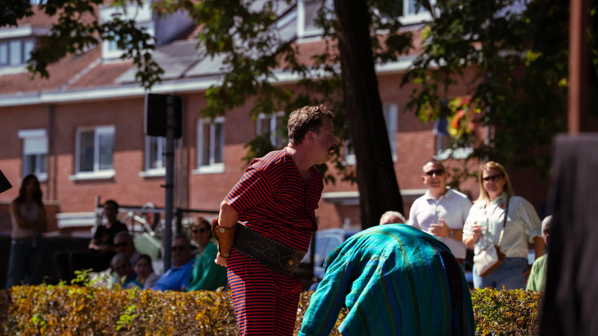 A man dressed in a red and black striped costume stands outdoors, crowd of onlookers watching, some smiling and wearing sunglasses, building with brick walls and windows in the background.