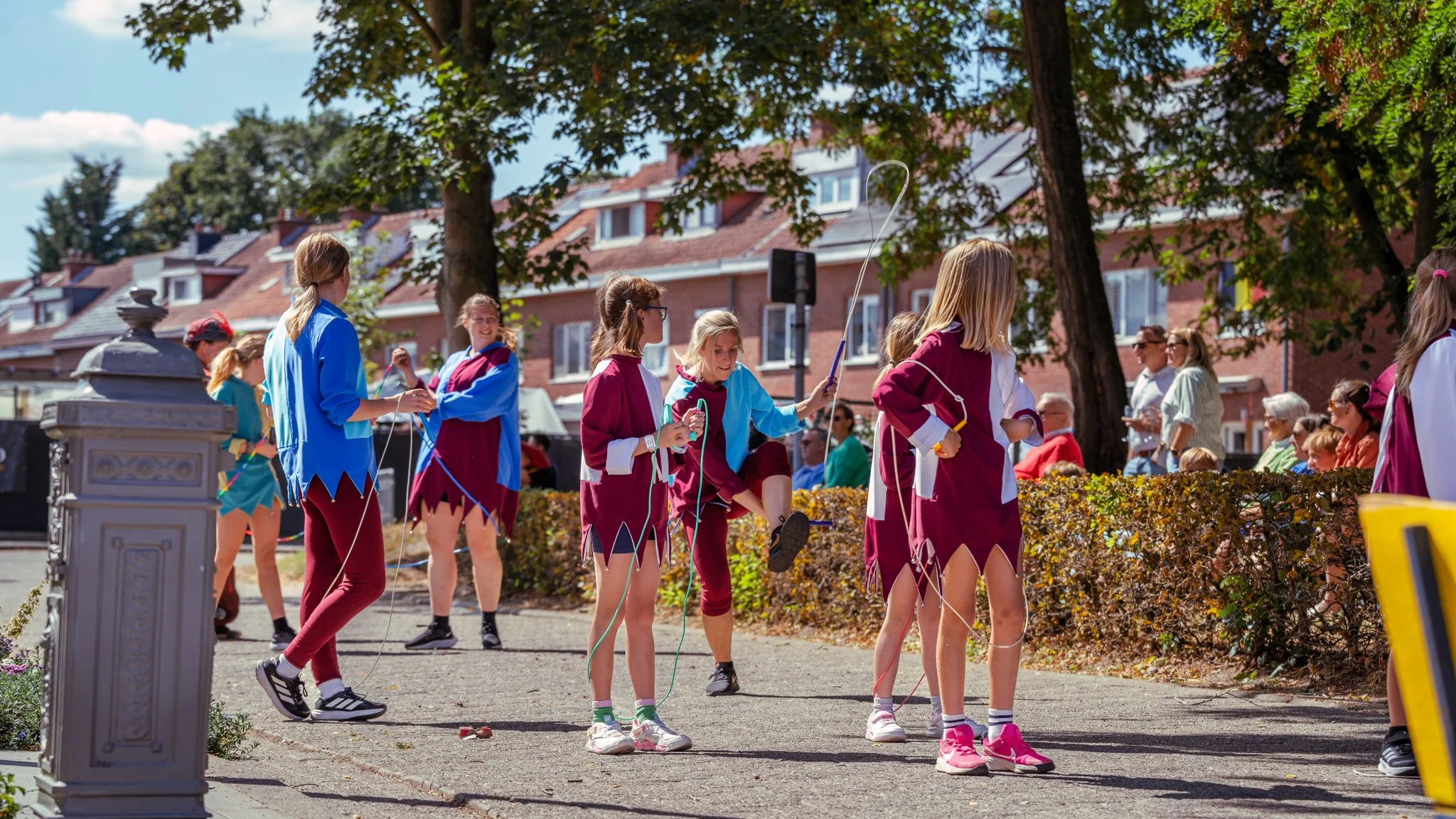 Children dressed in festive costumes holding balloons in a park during a parade, with spectators sitting on benches and houses in the background.