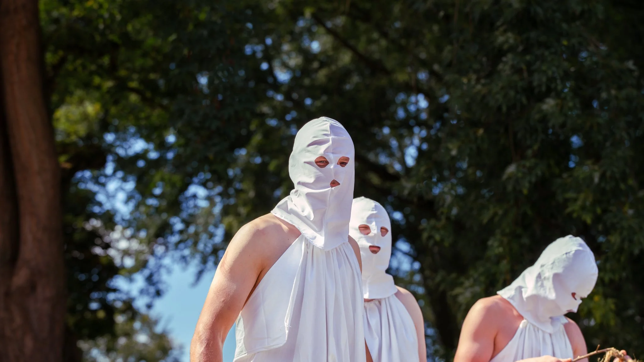 People wearing white masks with eye and mouth holes, dressed in white clothing, standing outdoors with a tree and blue sky in the background.