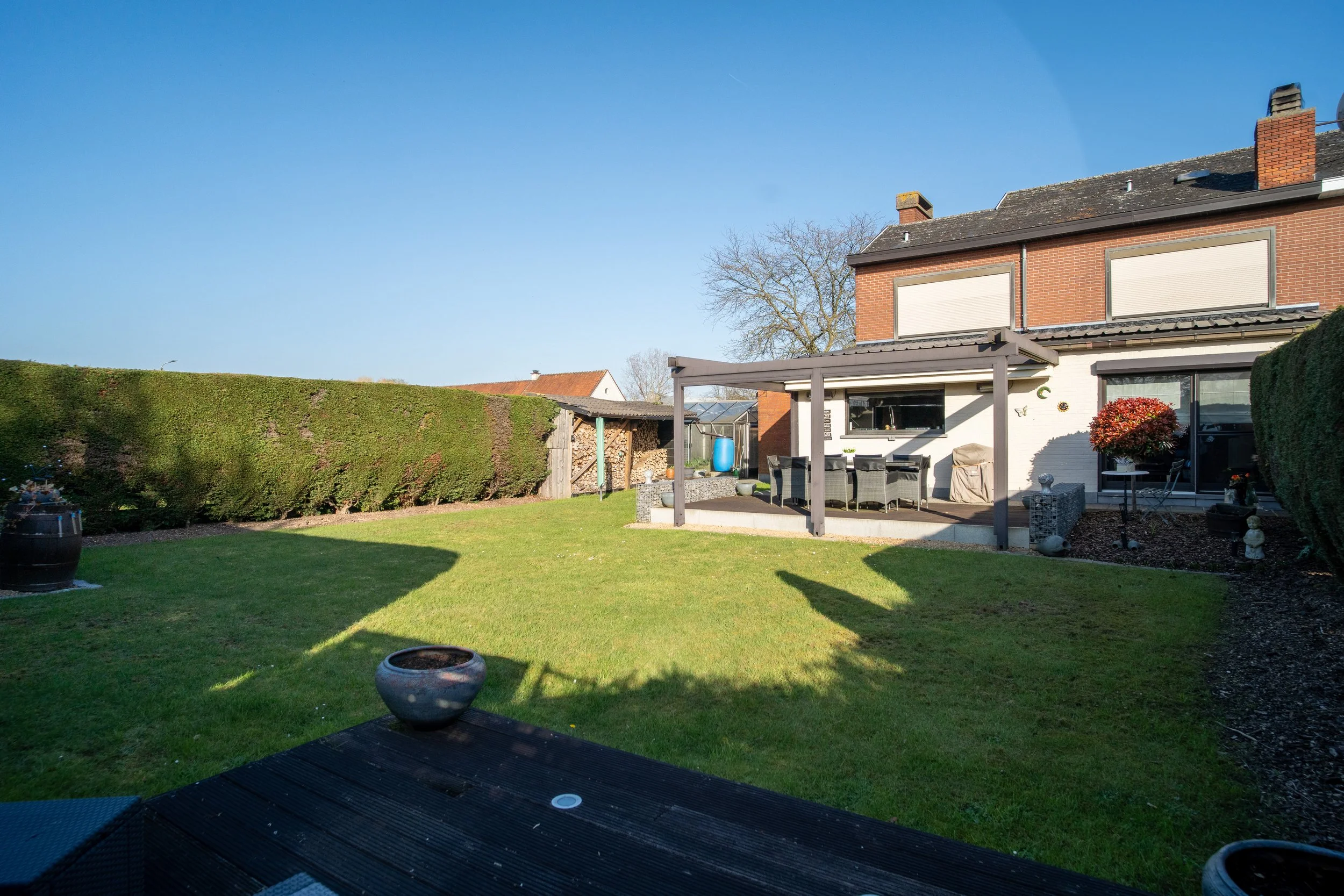 Backyard with a well-manicured lawn, large hedge, patio furniture, house with window shutters, blue sky, and a few distant trees.