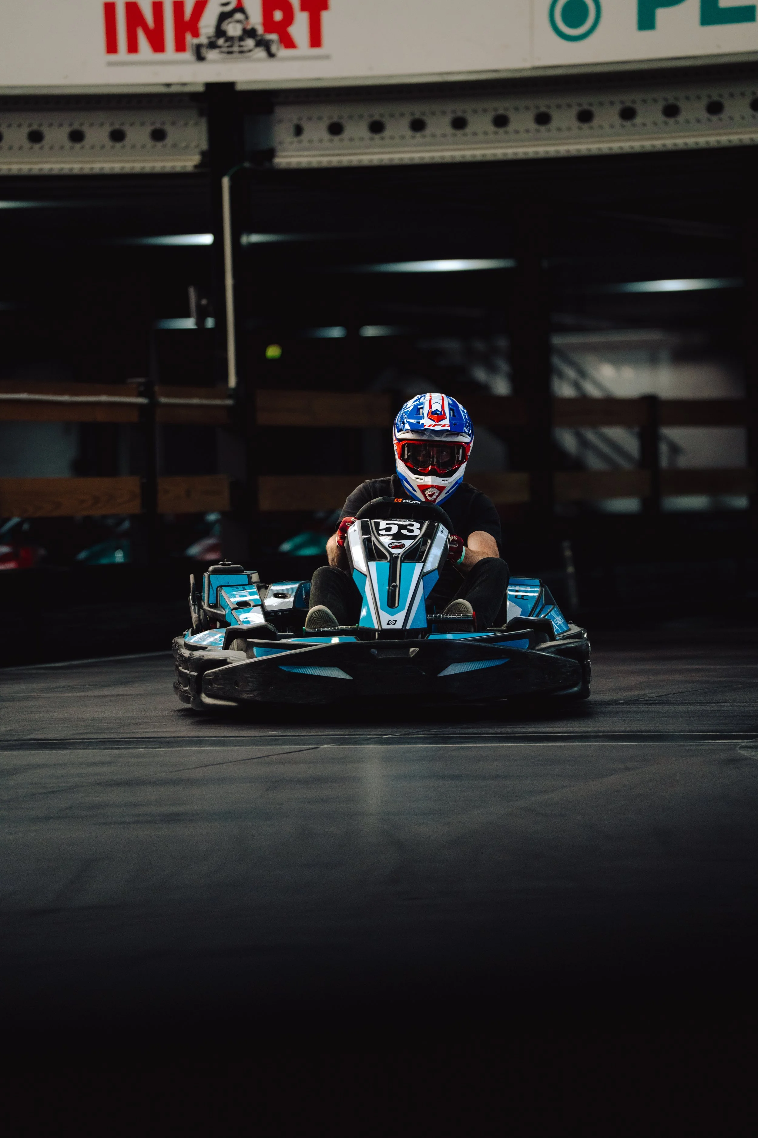 A person in a racing helmet driving a blue and white go-kart in an indoor racing facility.