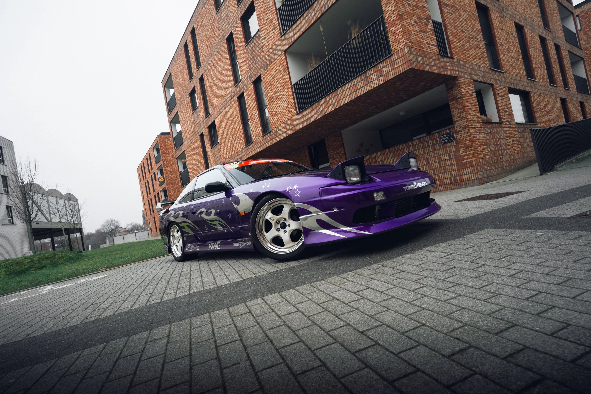 A purple sports car with racing decals parked on a brick driveway in front of modern apartment buildings.