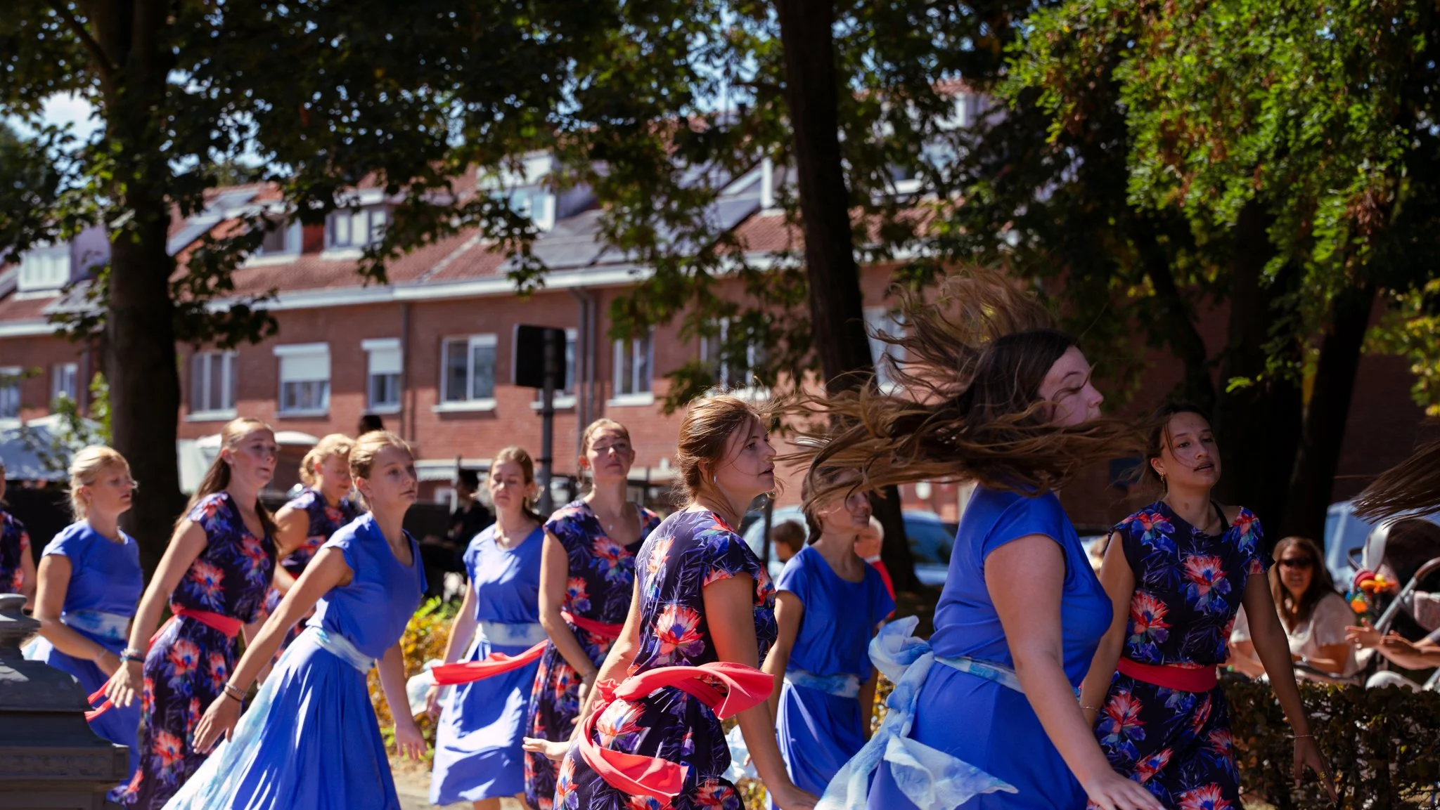 Group of young women in blue and floral dresses participating in an outdoor dance performance on a sunny day.