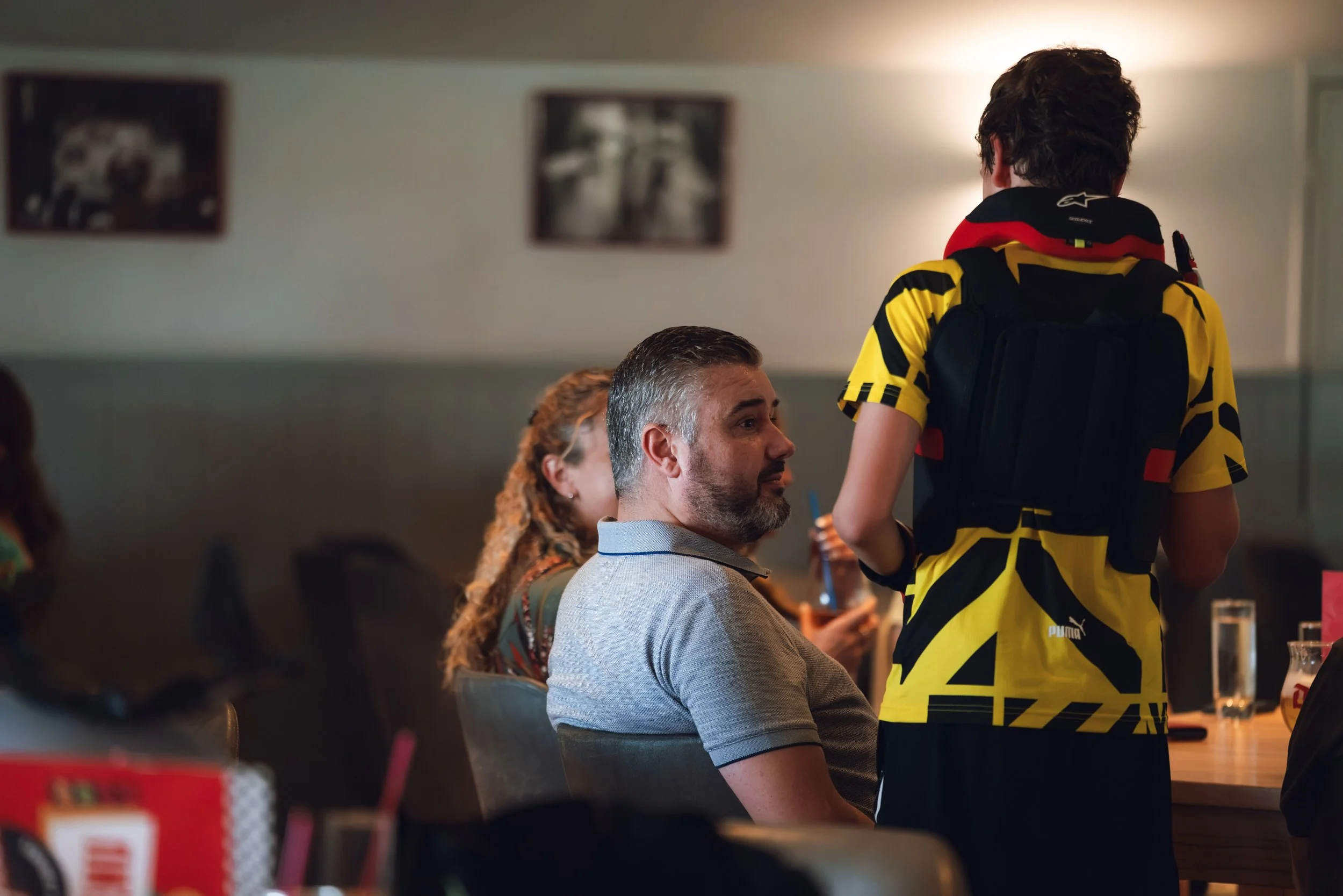 A man with gray hair and a beard is seated at a table, looking towards a person standing in front of him wearing a yellow and black sports jersey with a backpack. Several people are visible in the background, sitting at tables in what appears to be a