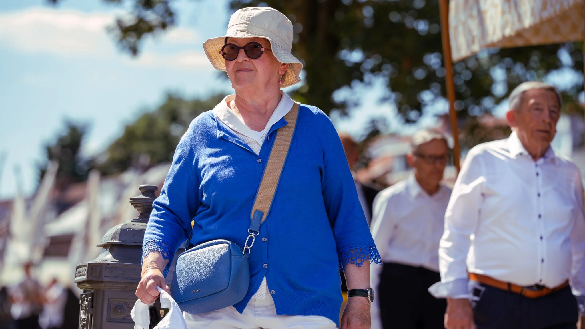 An older woman wearing a white hat, sunglasses, a blue cardigan, and white pants, walking outdoors. She has a brown crossbody bag and is holding a plastic bag in her right hand. There are other people in the background.
