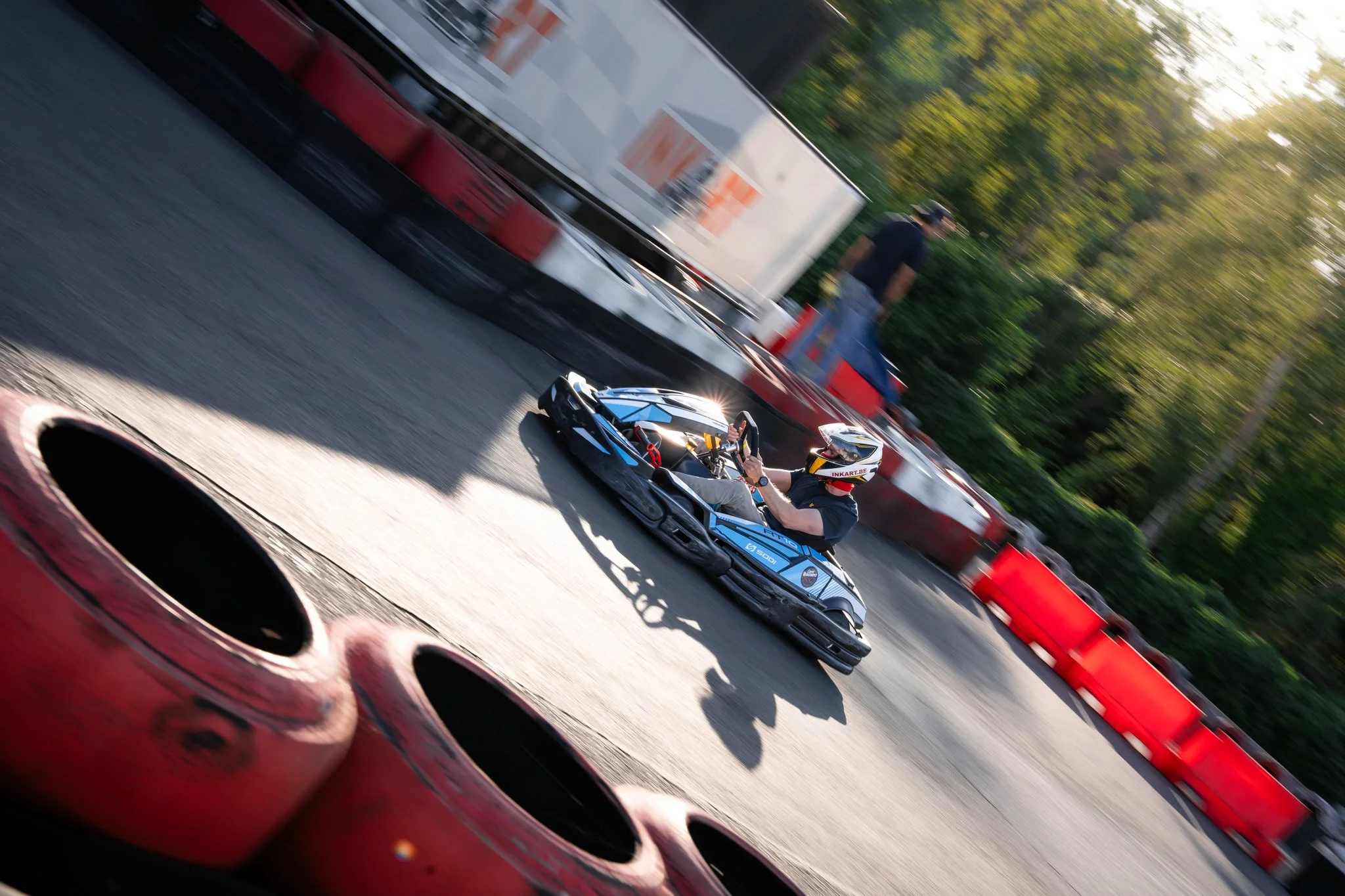 A person wearing a helmet driving a go-kart on an outdoor racing track with red barriers and trees in the background.