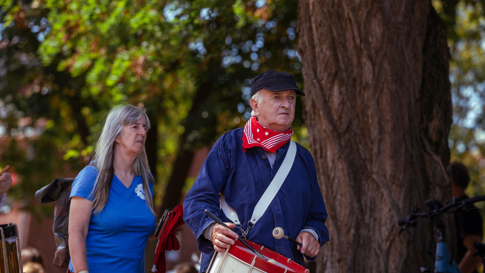 An elderly man with a red bandana around his neck, wearing a dark cap and blue jacket, is holding a drumstick and a small drum. Beside him, an elderly woman with long gray hair, wearing a blue shirt, looks on. They are outdoors near a large tree with