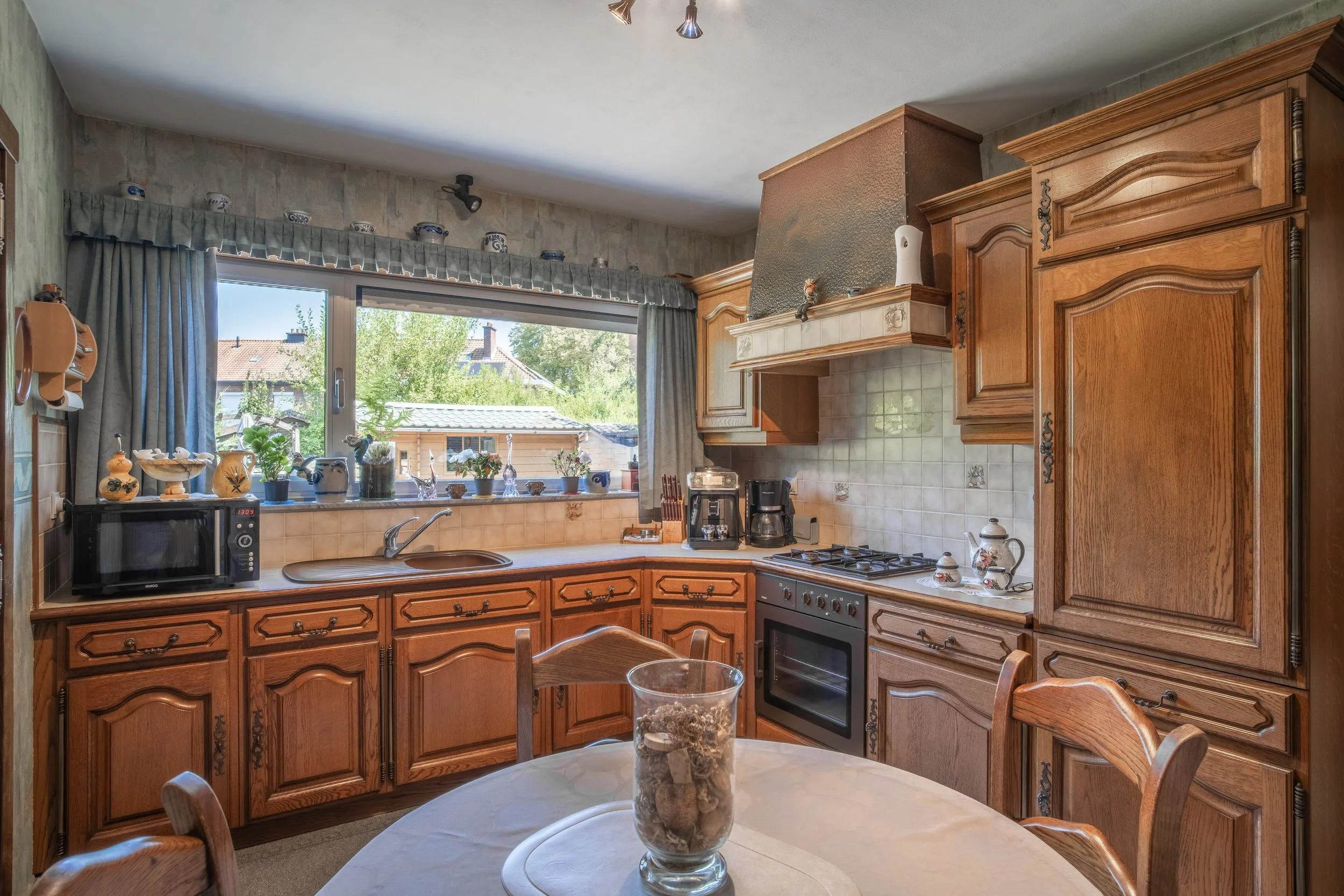 A kitchen with wooden cabinetry, a window with a view of trees and rooftops, and a table with chairs. There are decorative items on the window sill, a microwave, coffee maker, and stove visible.