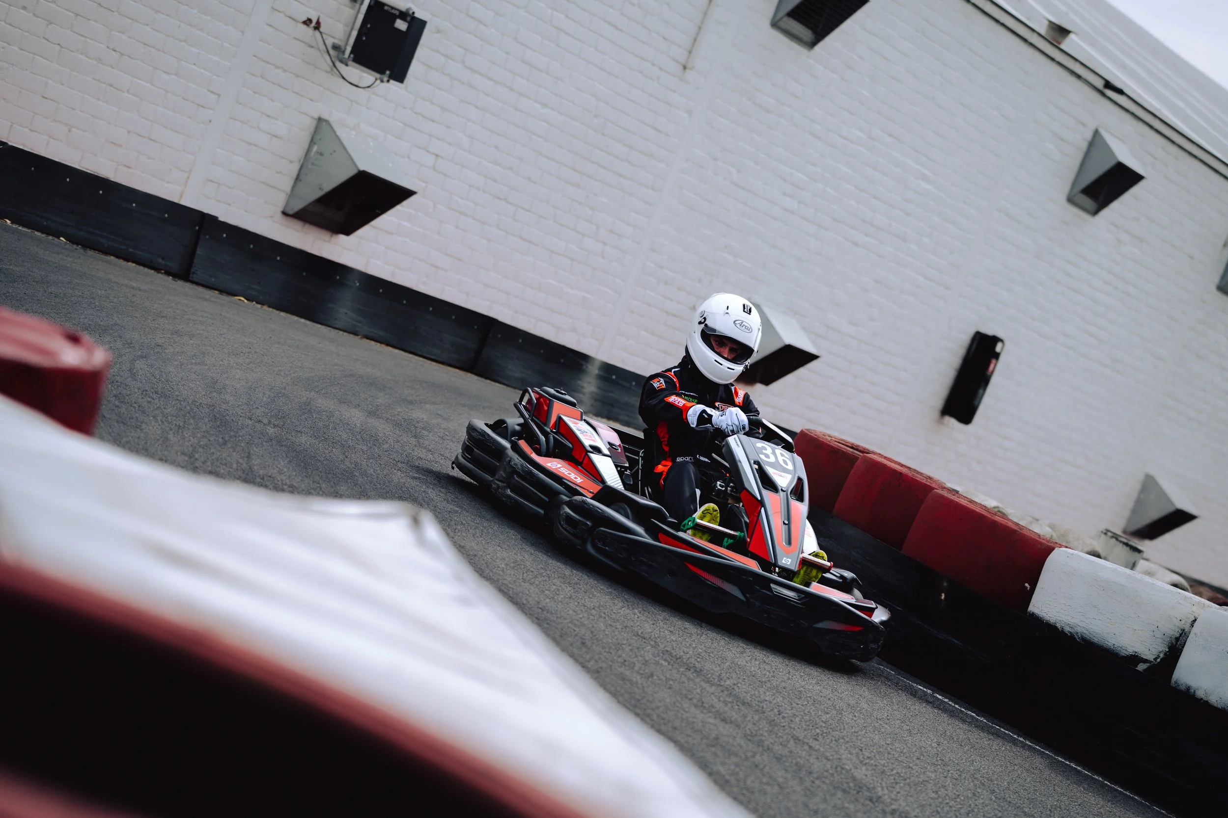 A person in racing gear and a white helmet driving a go-kart on a race track, with a white brick building and red barriers in the background.