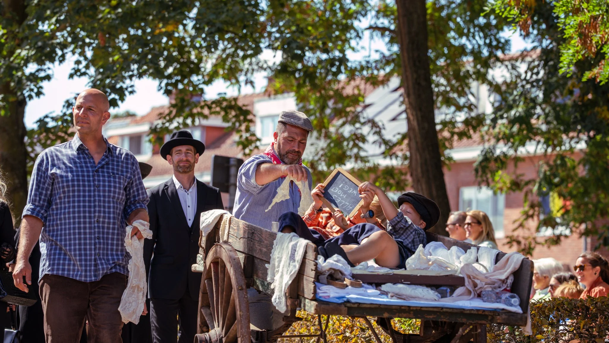 People dressed in vintage clothing gathering outdoors, with some sitting in a wooden cart surrounded by trees and older buildings in the background.