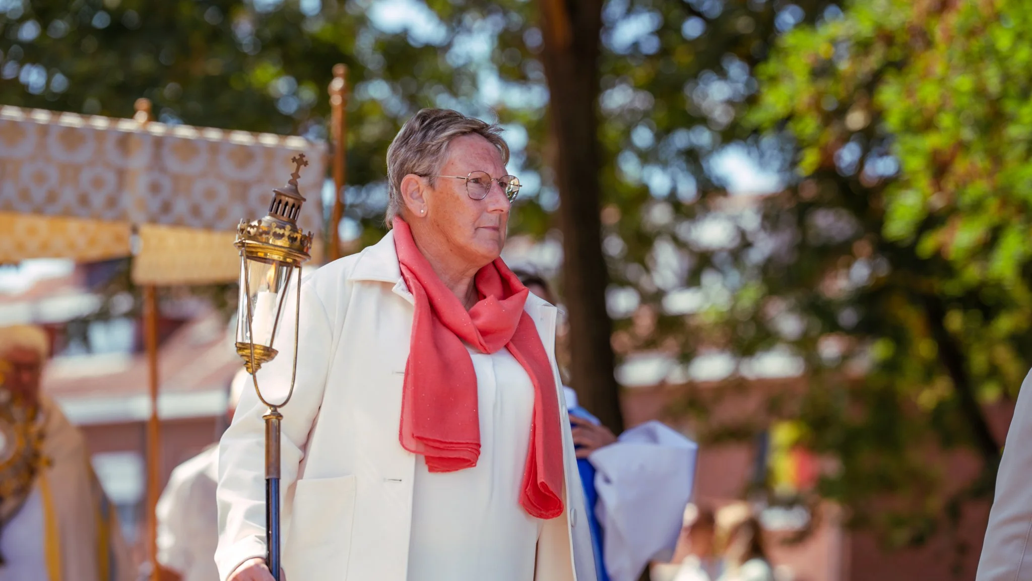 An elderly woman with short gray hair, glasses, wearing a white jacket and coral scarf, standing outdoors at a procession holding a lantern.