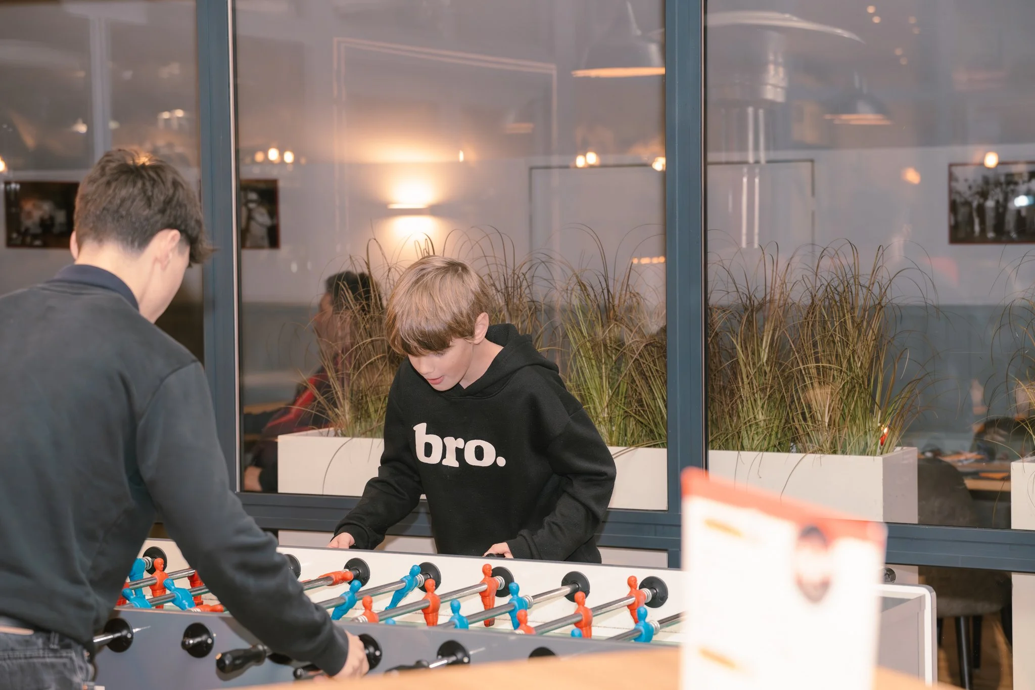Two boys playing foosball game inside a room with large windows and a plant.