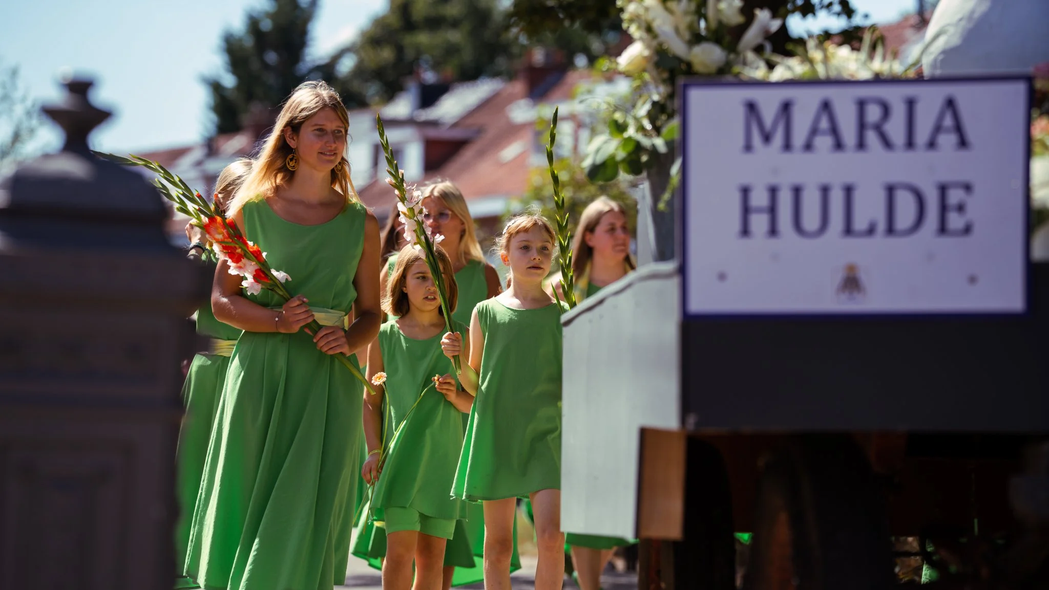Group of women and girls wearing matching green dresses participating in a procession, holding gladiolus flowers, walking on a sunny street near a decorated float with a sign that reads 'Maria Hulde'.