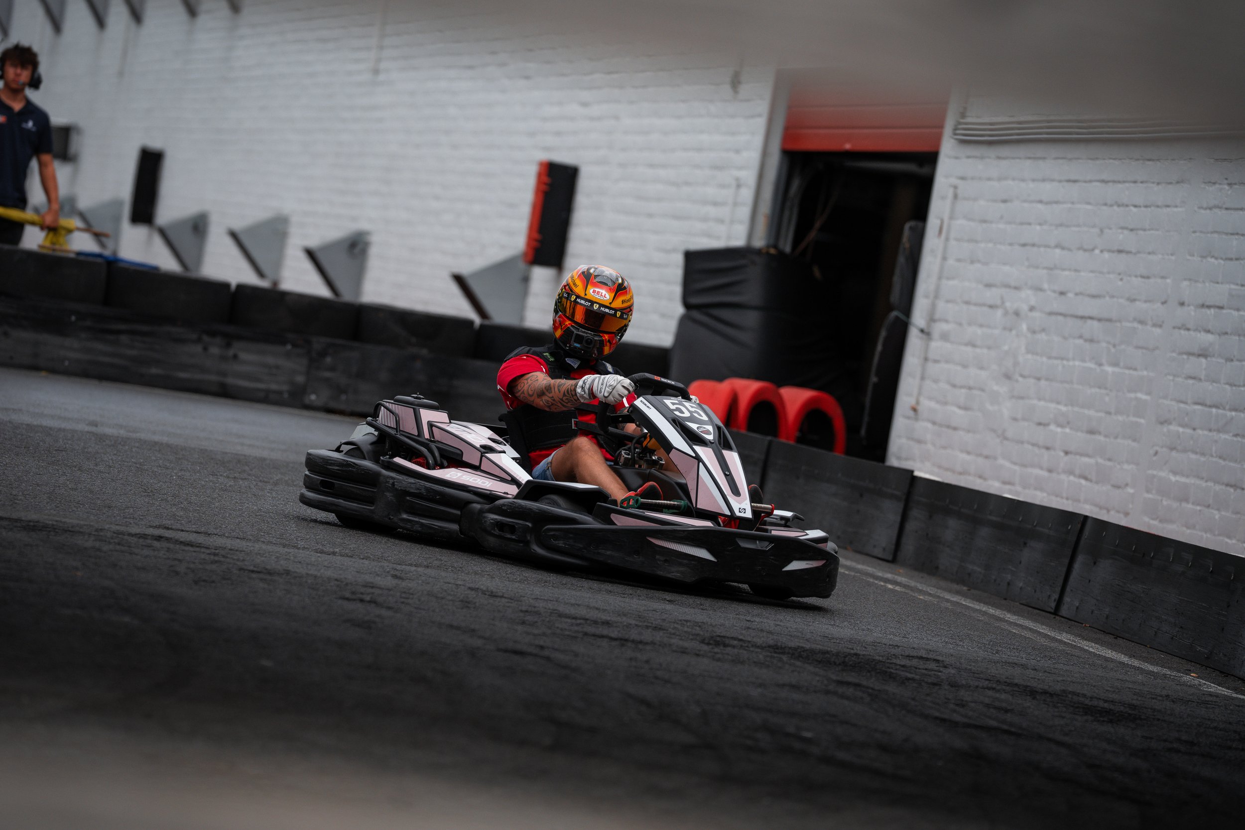 A person wearing a motorcycle helmet and red shirt driving a go-kart on an indoor racing track with a white brick wall background.