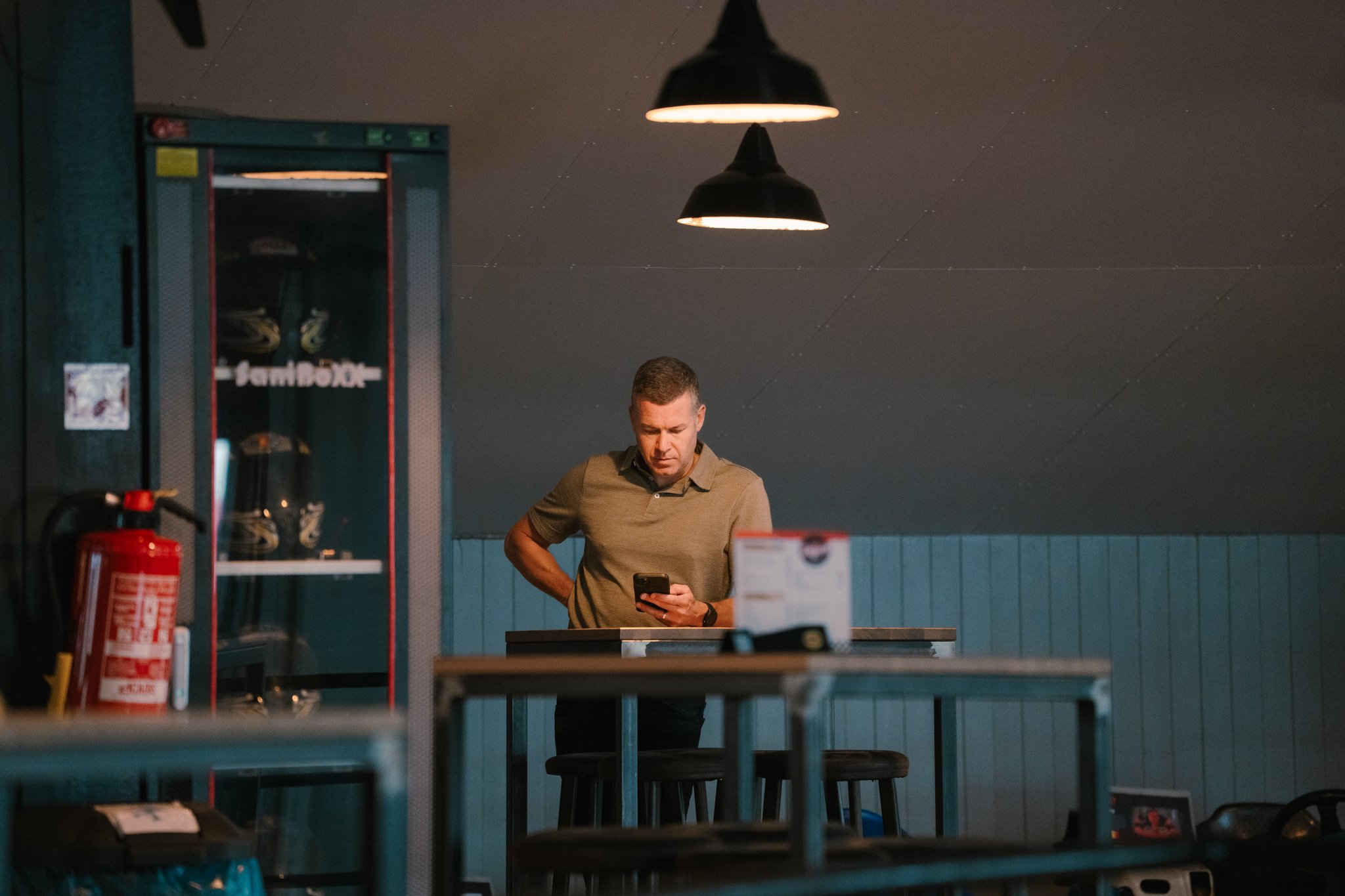 A man is standing at a high table in a dimly lit room, looking at his phone. There are black pendant lights hanging above him, and a fire extinguisher is visible on the left side of the image. The background features a dark ceiling and teal paneling 