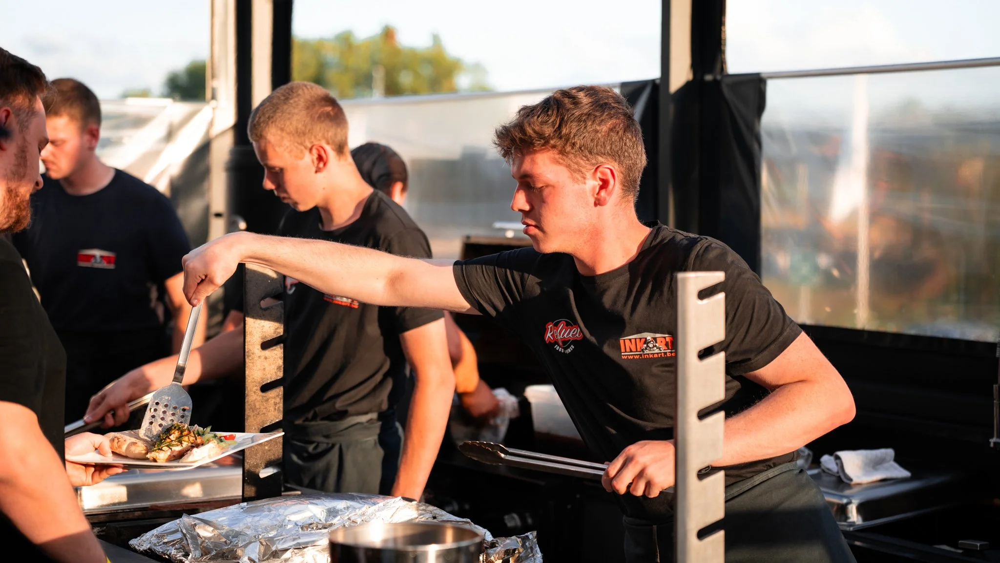 A group of young men working at a food stand, serving food to a customer in an outdoor setting with a tent and trees in the background.