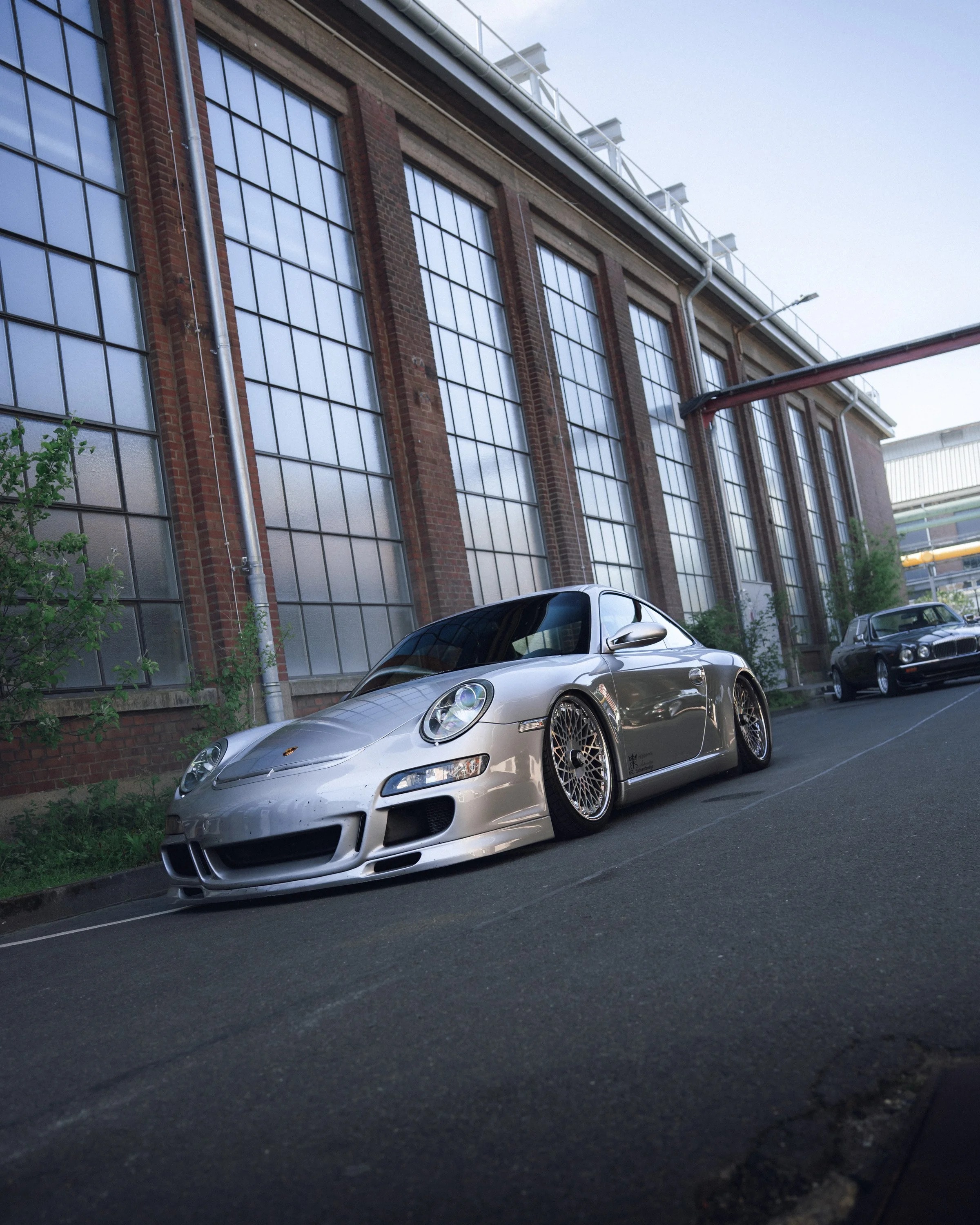 A silver Porsche sports car parked on the street in front of a building with large windows and brick walls.