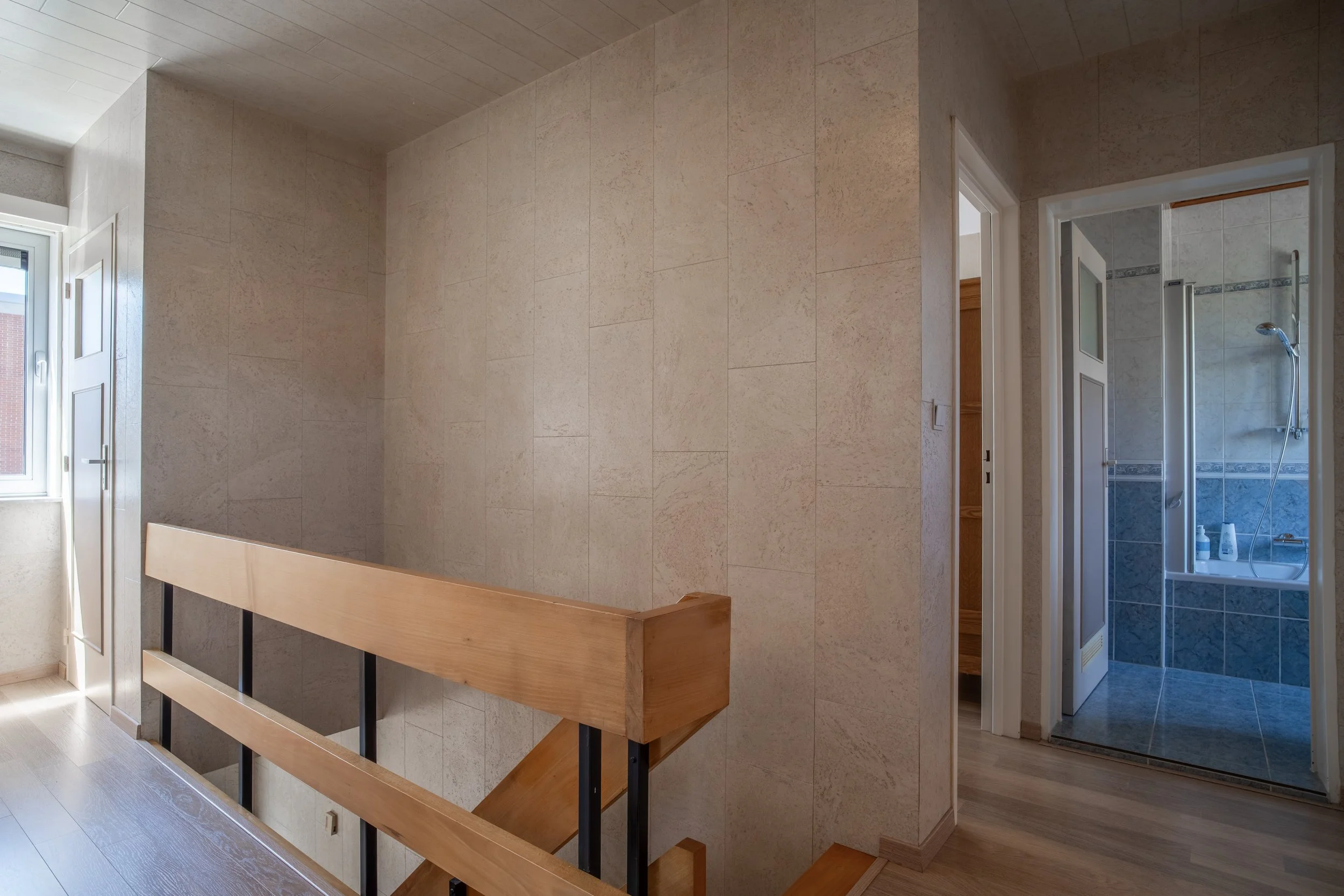 Interior view of a modern home with beige tiled walls, wooden floor, a staircase with wooden handrails, and a bathroom with a shower visible through a door.