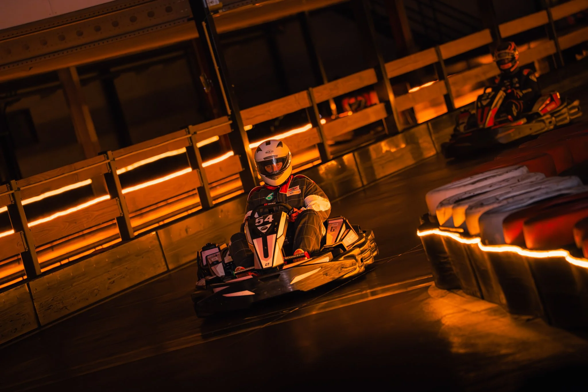 Two race car drivers in full gear racing on an indoor track with warm orange lighting, one in the front with number 54 and another in the background.