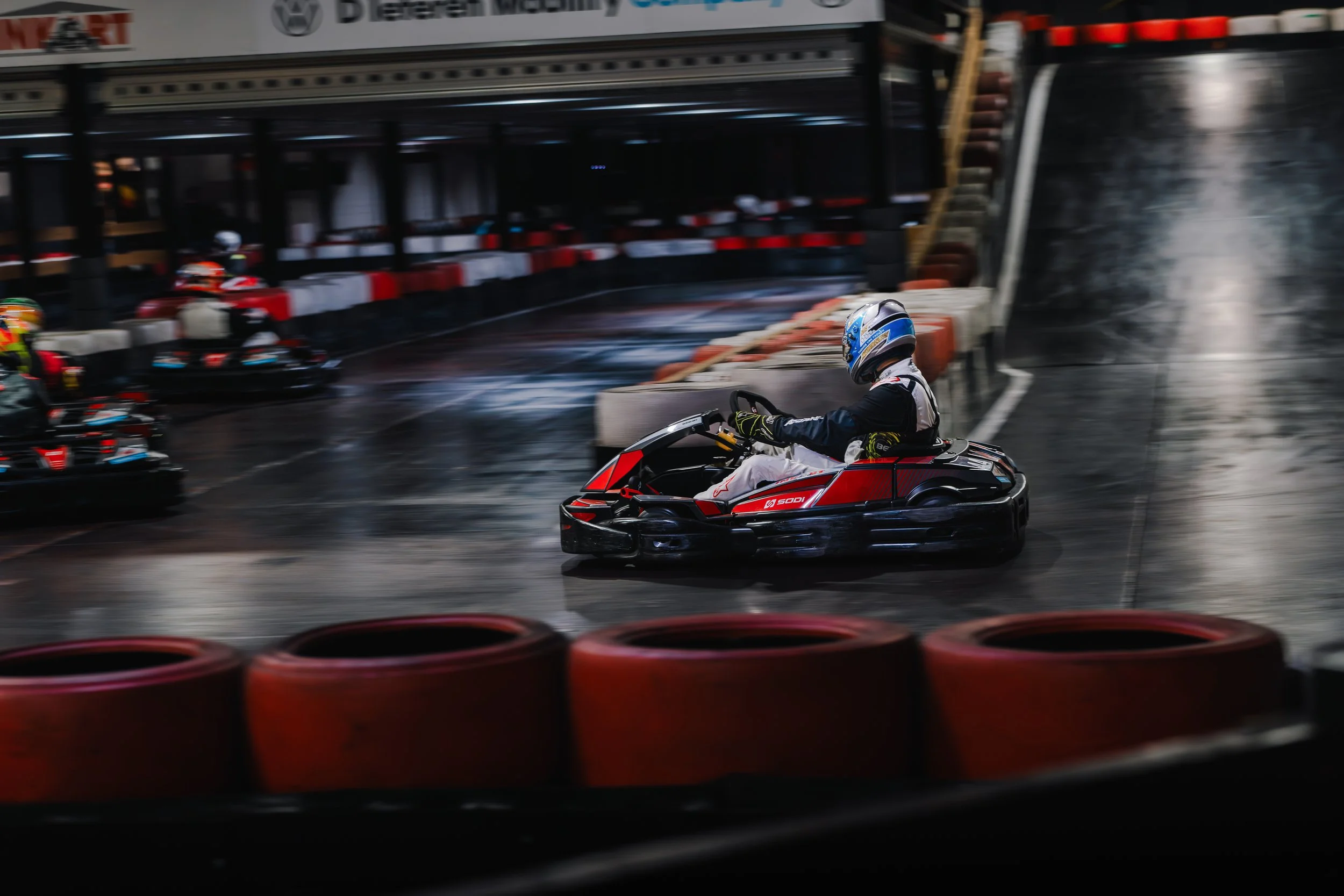 A person wearing racing gear and a helmet driving a go-kart on an indoor racing track with red and black barriers, other go-karts and racers visible in the background.