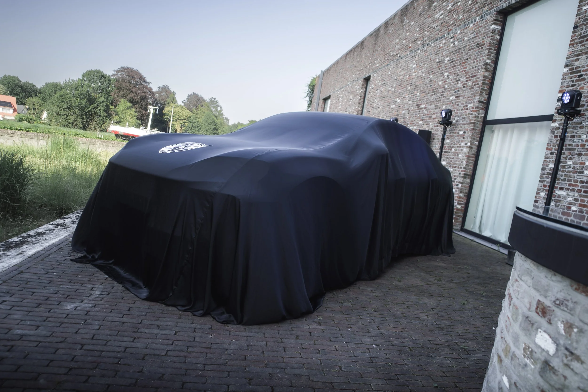 A covered object, possibly a car, parked outside near a brick building with two outdoor lights. The object is draped in a black cover with a logo. The background shows trees and a clear sky.