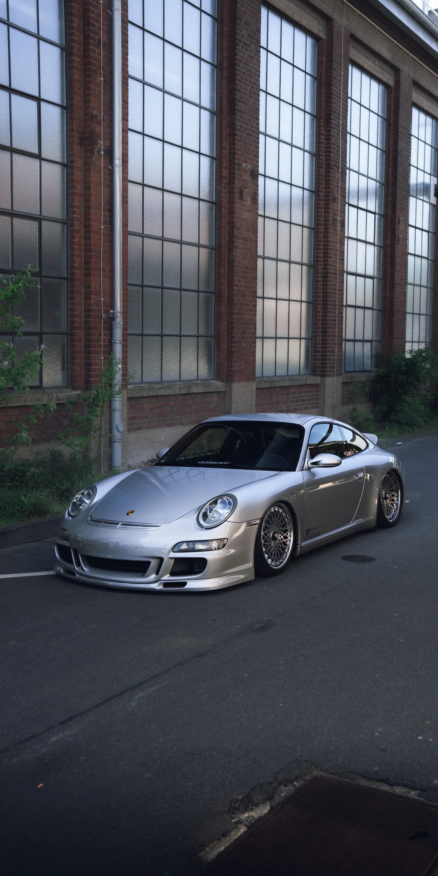 Silver Porsche sports car parked on city street with large industrial-style brick building with tall windows in the background.
