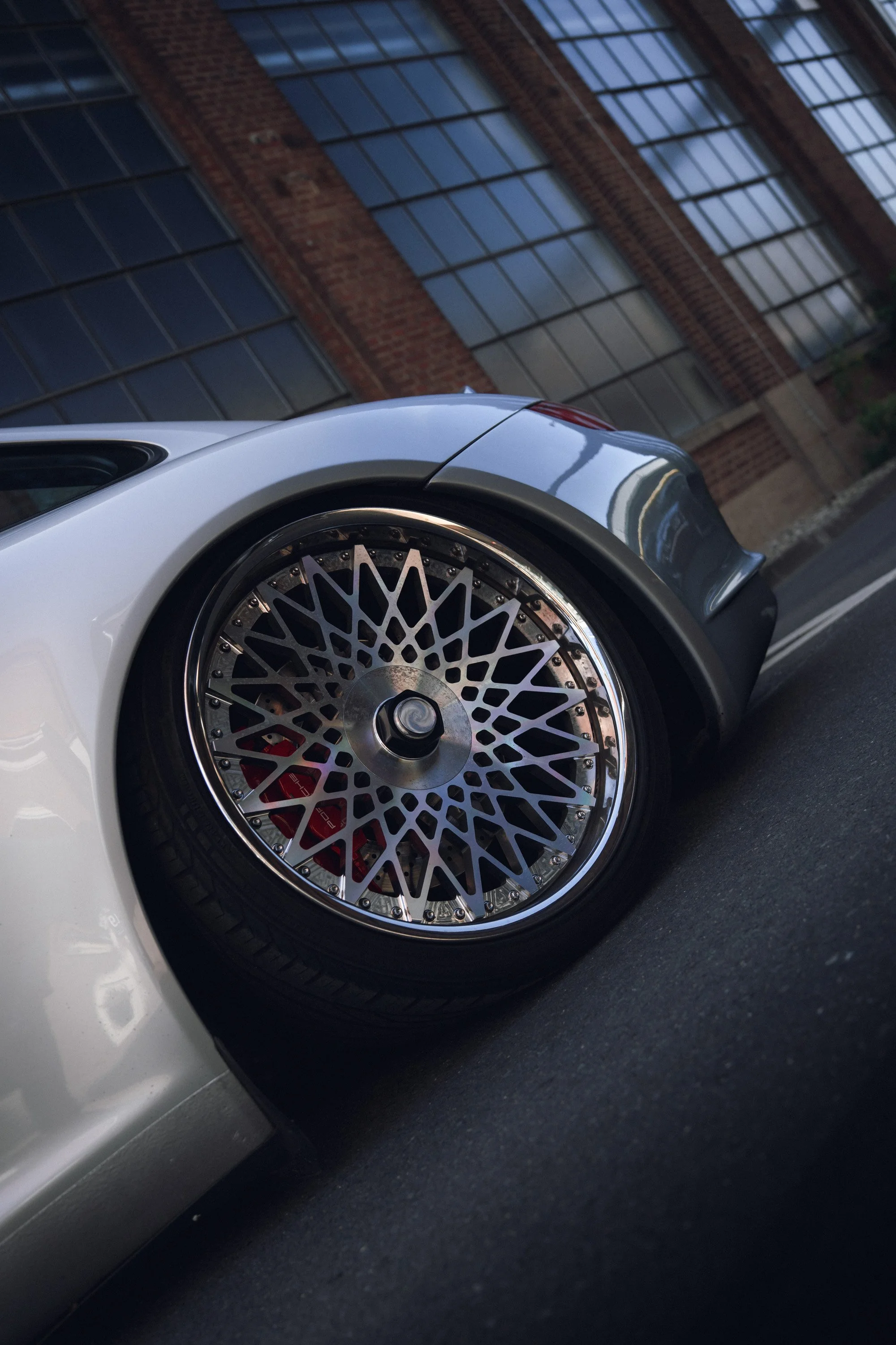 Close-up of a modern silver car with a detailed multi-spoke alloy wheel, parked on a street with a brick building and large pane windows in the background.