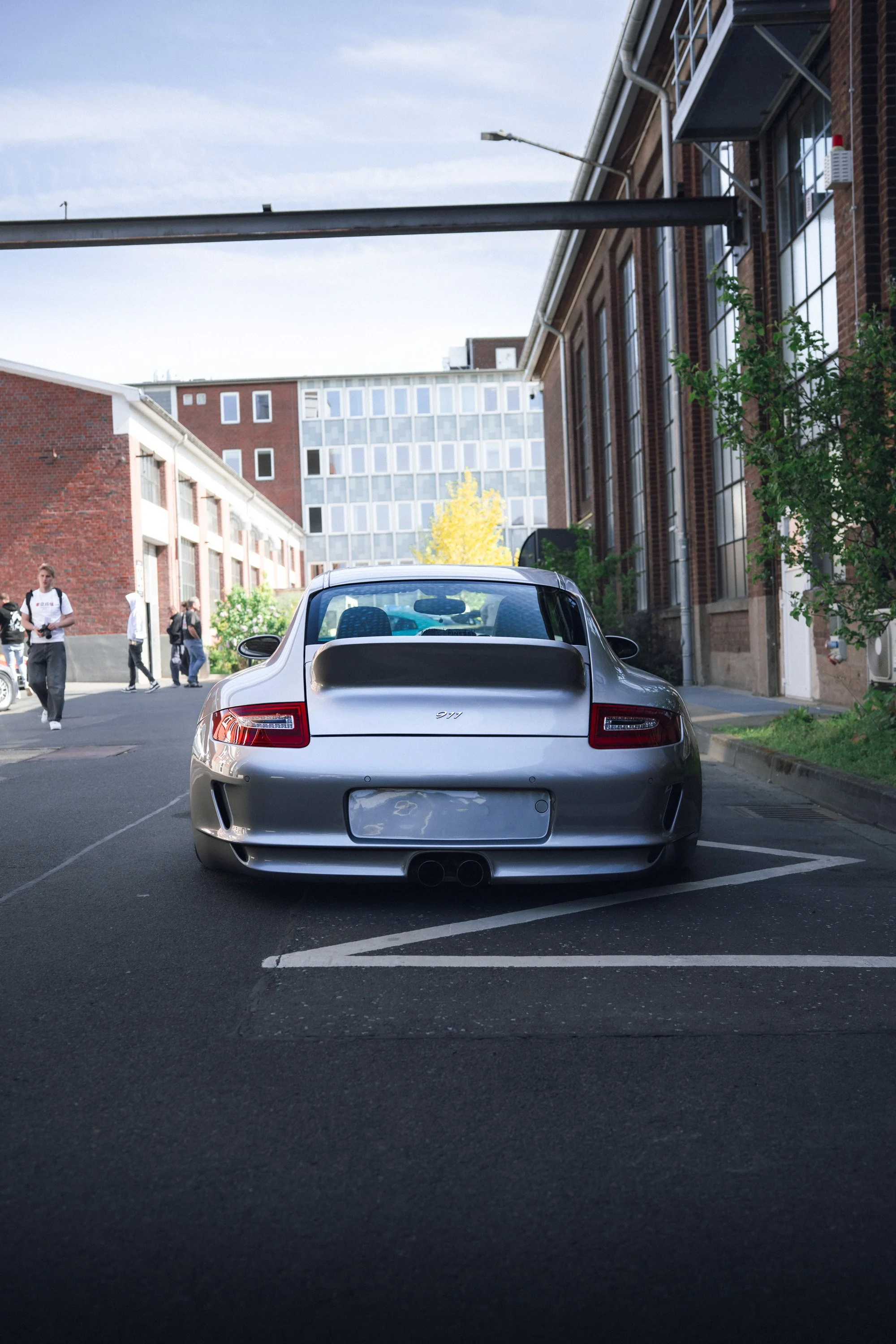 Silver Porsche parked in an outdoor parking lot with buildings and pedestrians in the background.