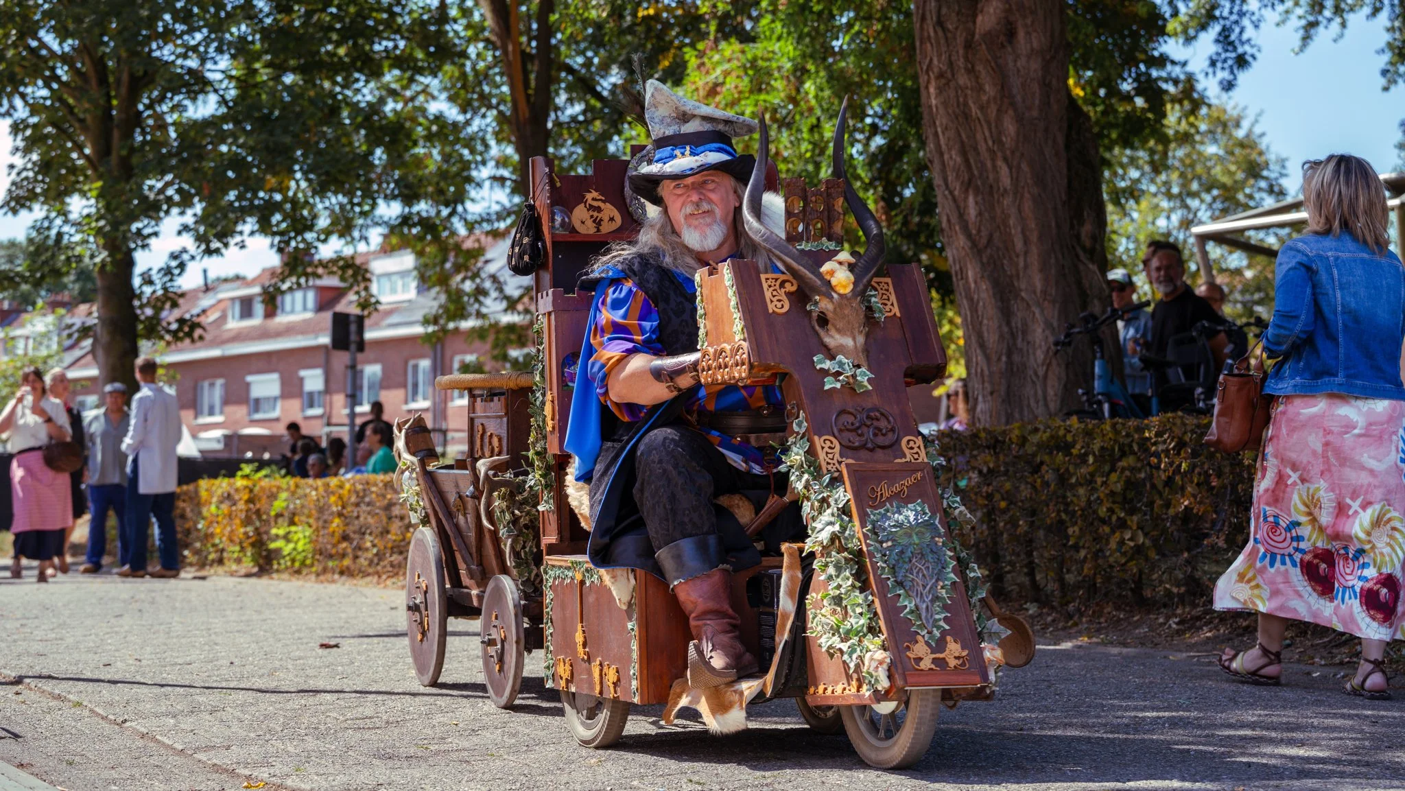 A man dressed as a wizard rides a decorated wooden cart during a festival or parade in an outdoor park area, with people and trees in the background.