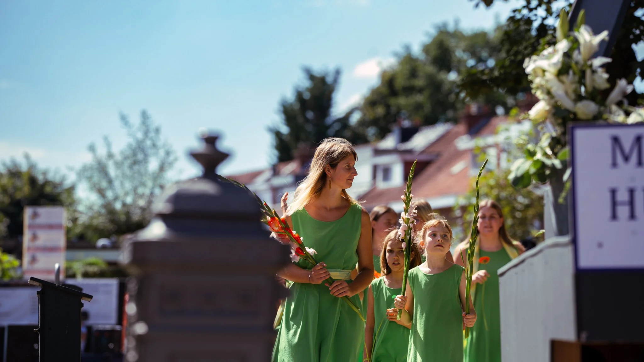 Women and girls dressed in green gowns holding flowers participate in a memorial event outdoors on a sunny day.