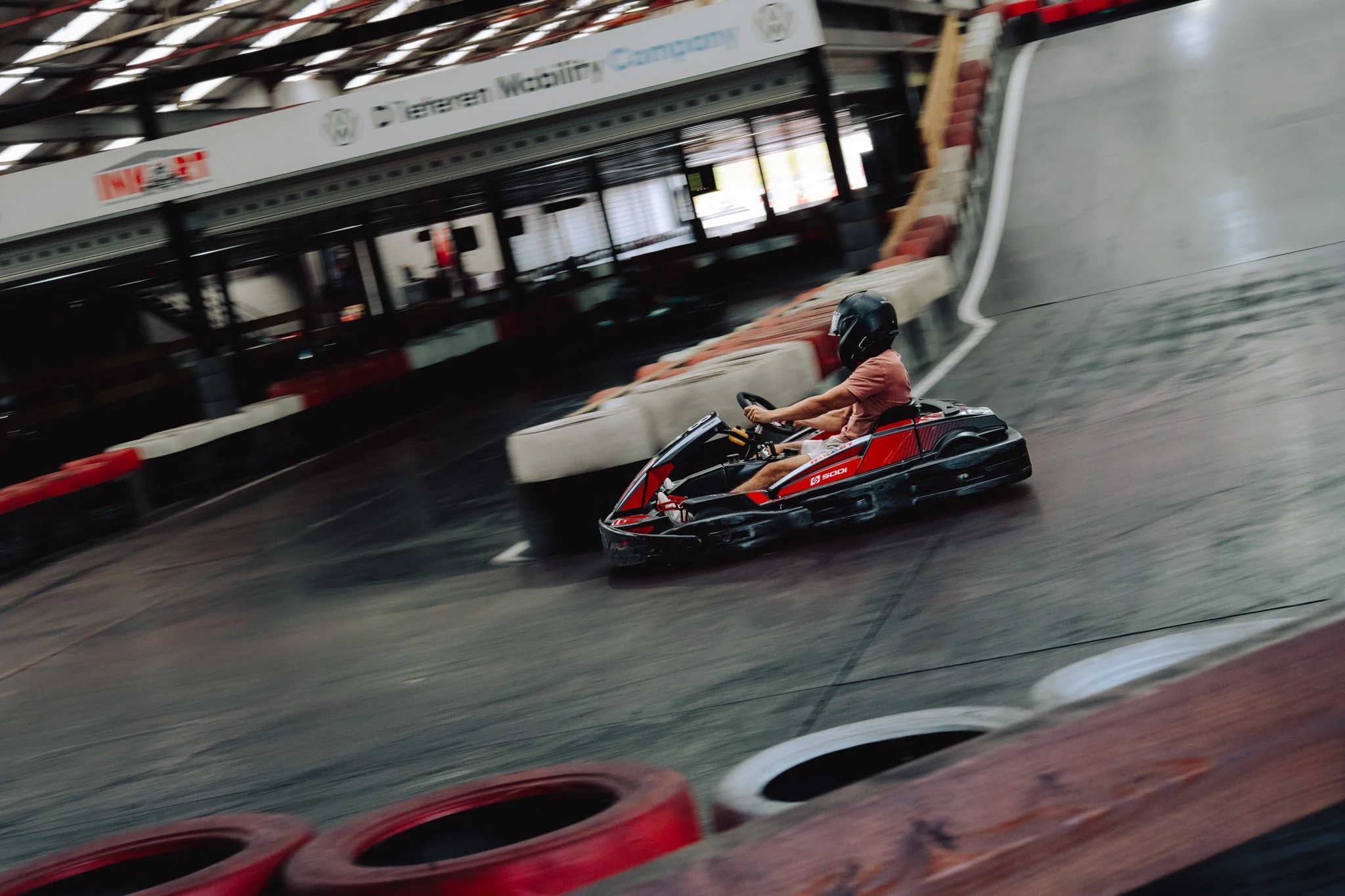 Person wearing a helmet driving a red and black go-kart on an indoor race track.