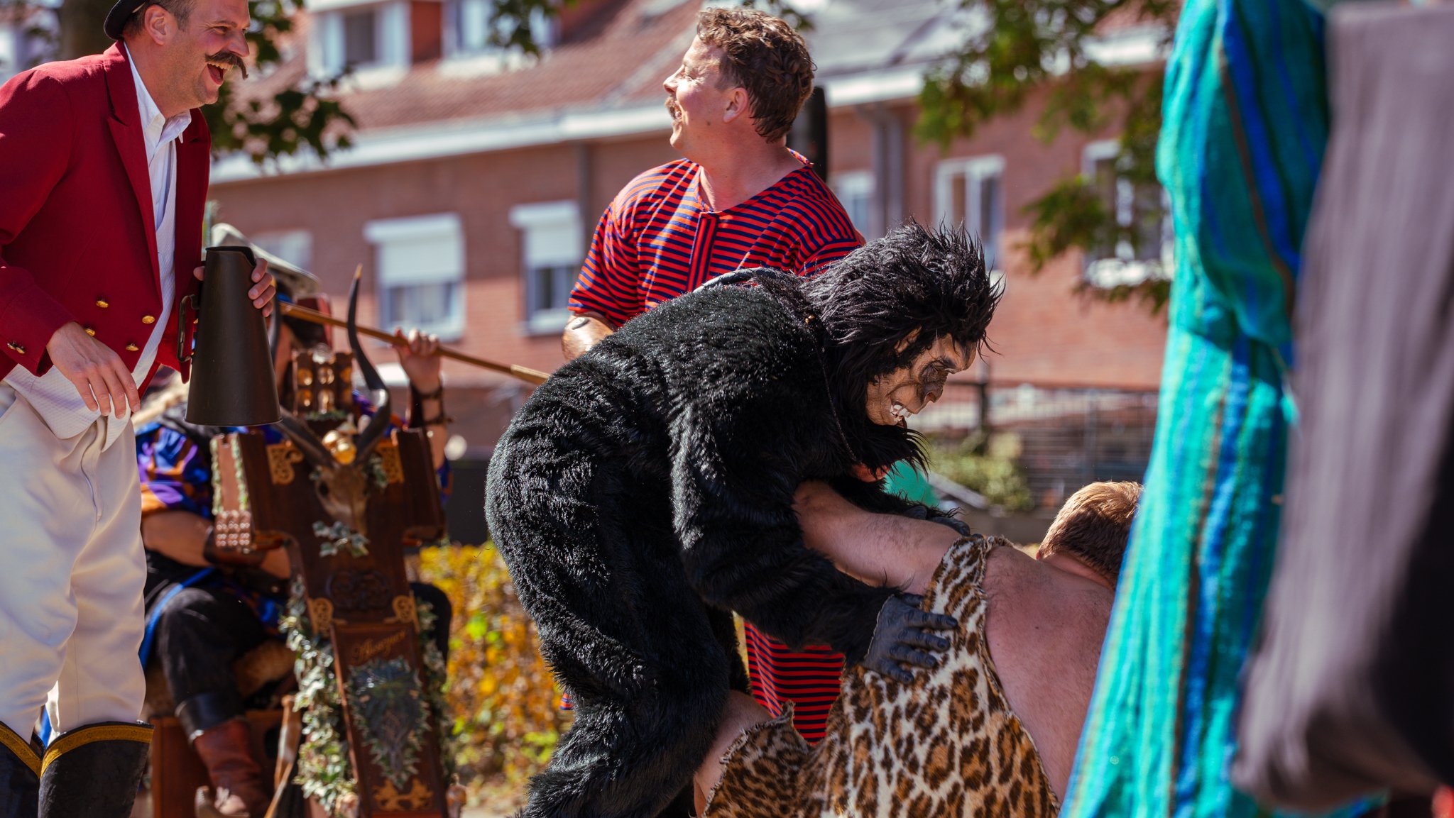 People dressed in costumes participating in a lively outdoor event or festival, with buildings and trees in the background.
