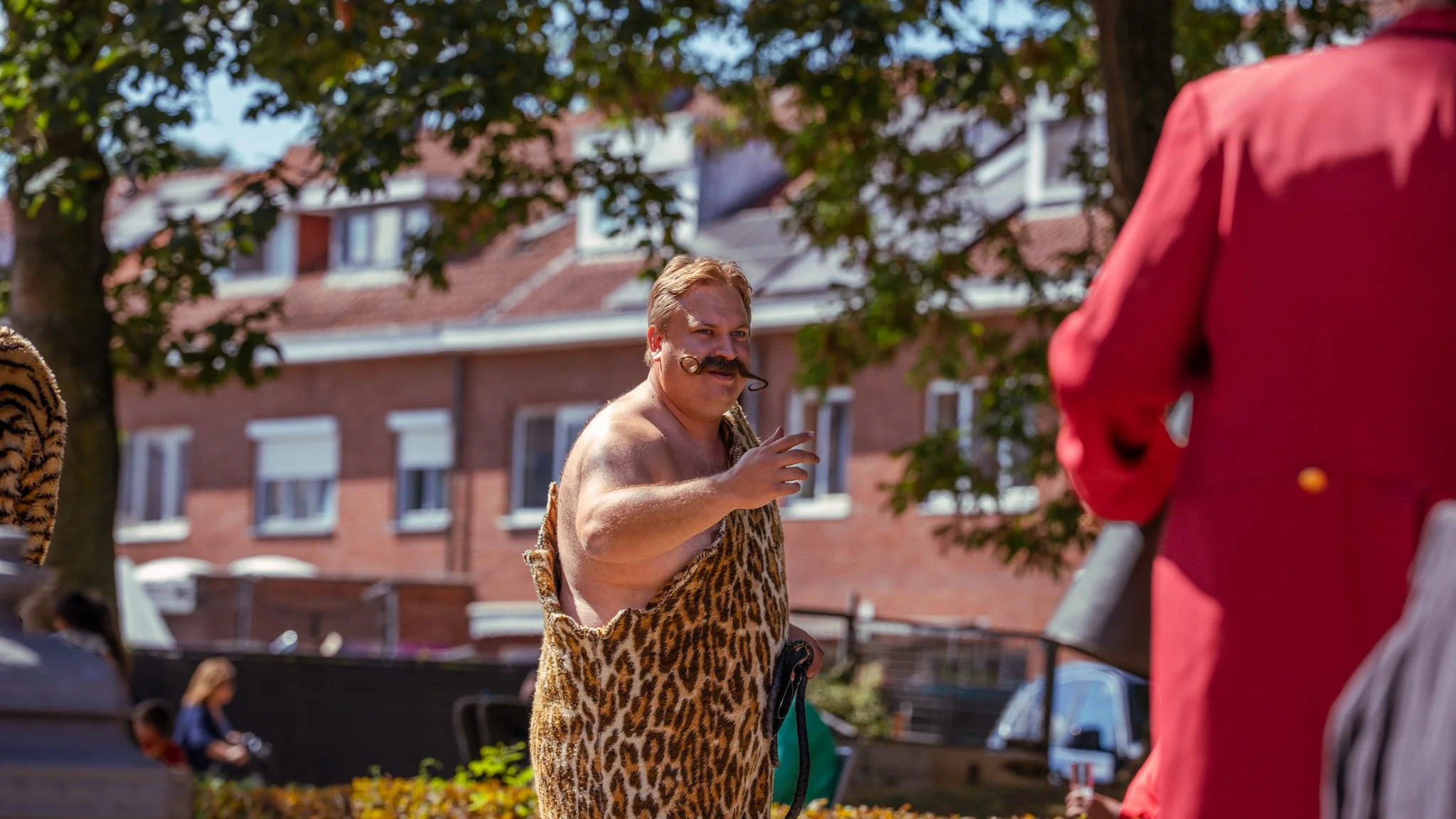 Man dressed in a leopard print costume with a mustache, standing outdoors in a neighborhood, gesturing with his fingers, with houses and trees in the background.
