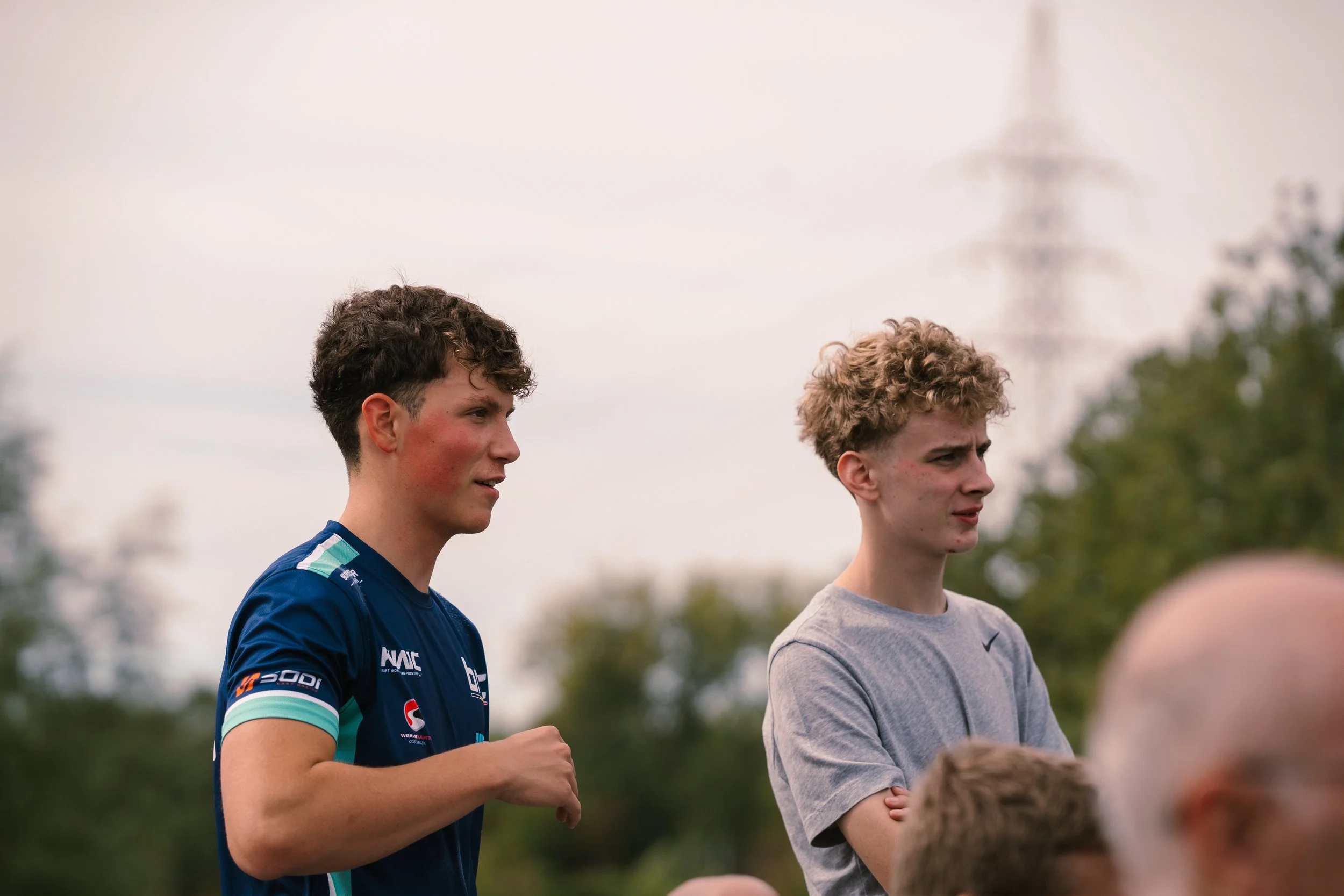 Two young men standing outdoors, looking focused, with trees and a cloudy sky in the background.