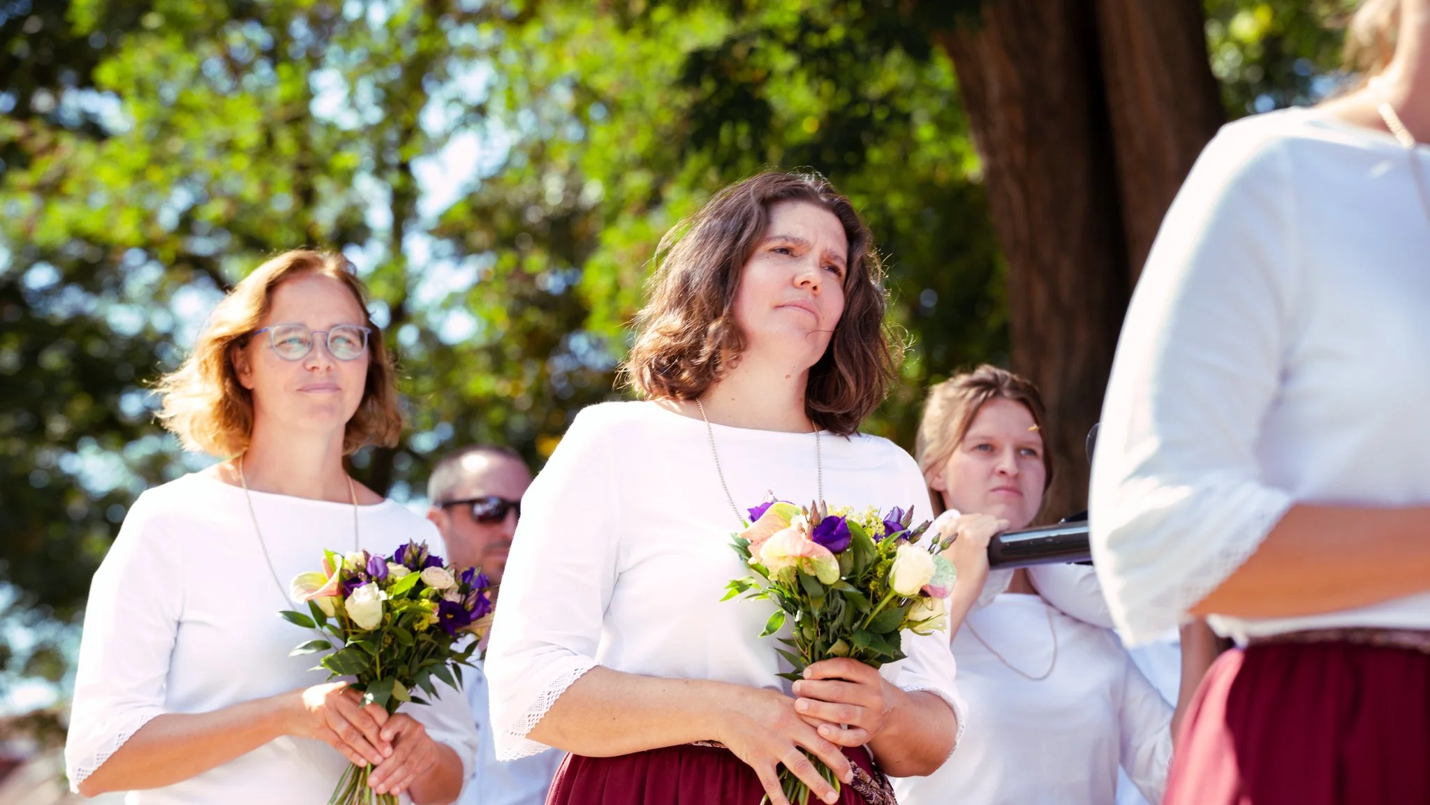 Women standing outdoors holding bouquets of flowers during a ceremony, with trees in the background.