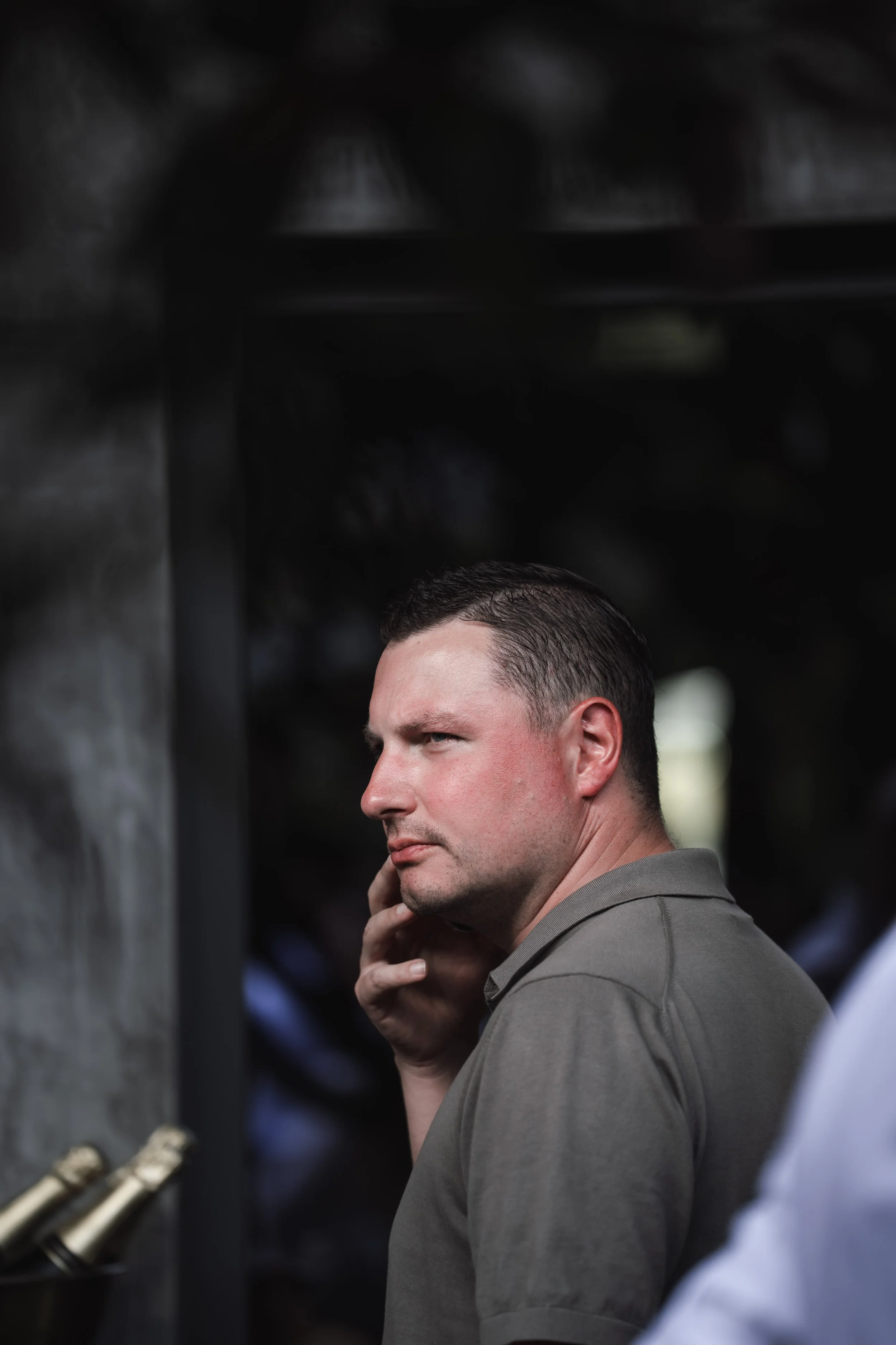 A man with short dark hair and a grey shirt is talking on a cell phone with a serious expression, standing outdoors near a dark structure.