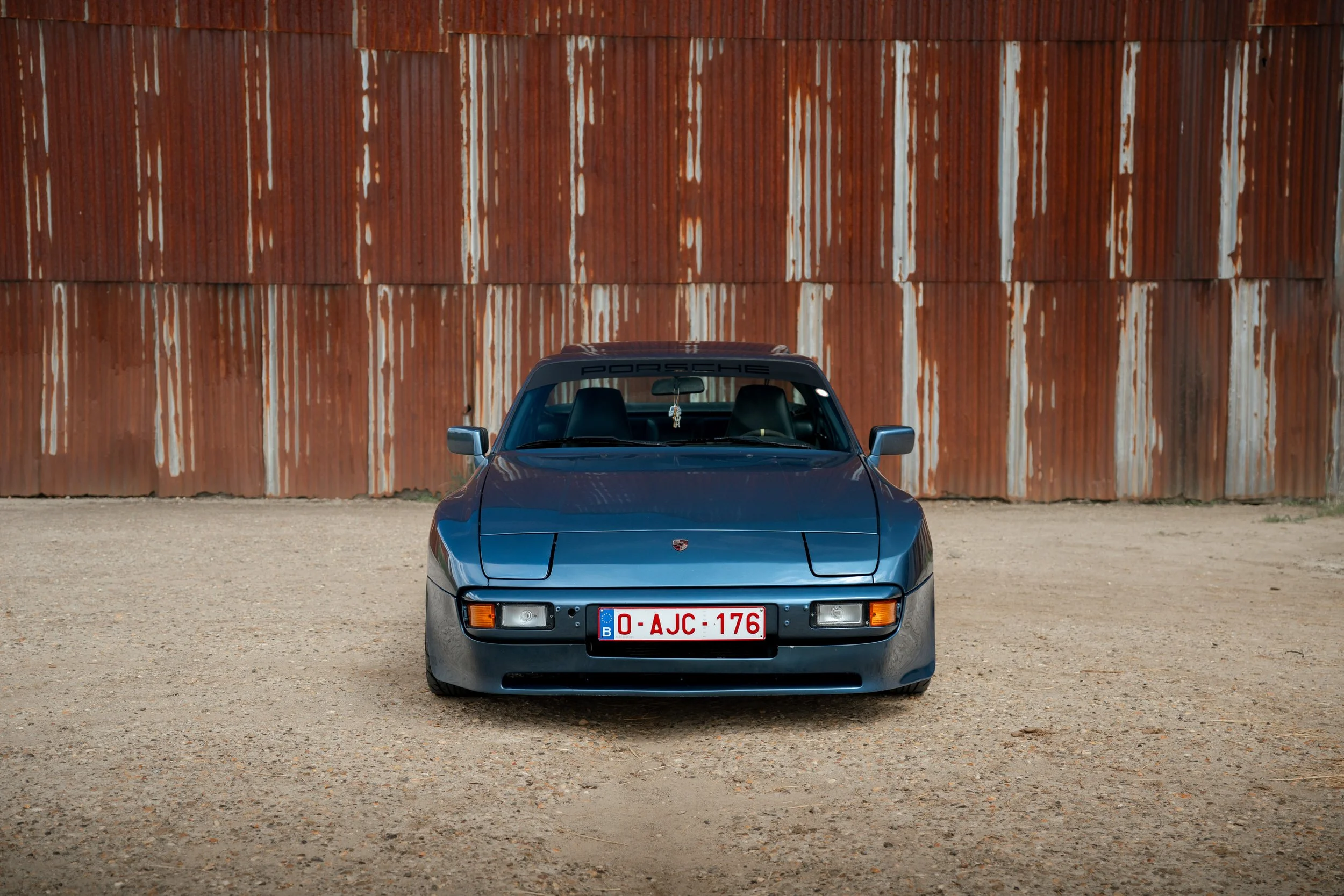 A vintage blue Porsche car parked in front of a rusted metal wall.