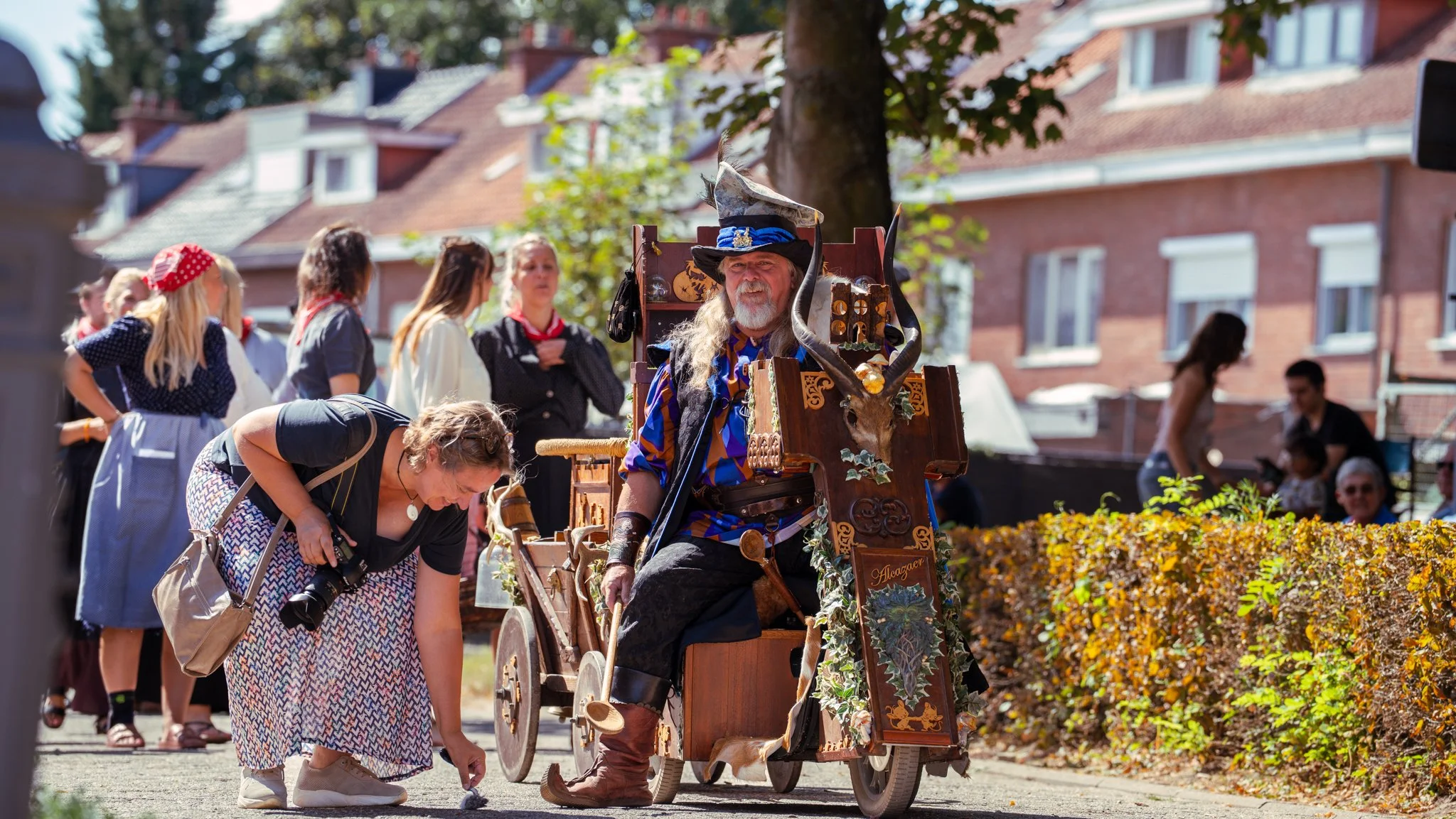 A man dressed as a wizard, seated in a wooden cart decorated with fantasy-themed items, at an outdoor event. The wizard wears a tall pointed hat, long hair, and a robe, and has a long gray beard. Several people are gathered around, some dressed in co