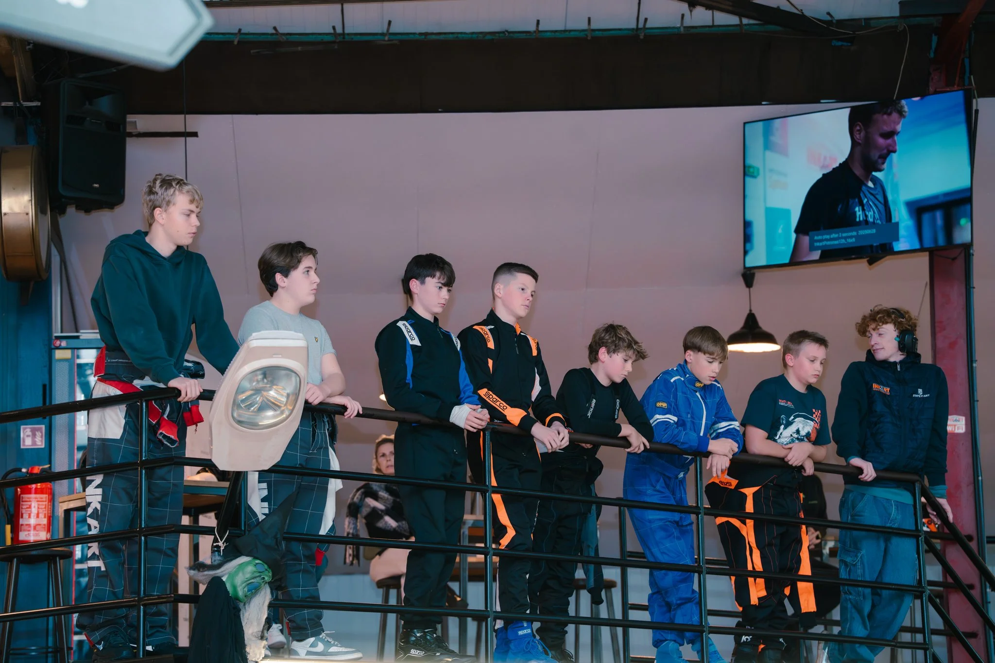 Group of young boys in racing suits standing behind a railing, watching an event in an indoor racing facility with a large TV screen showing a man in racing gear.