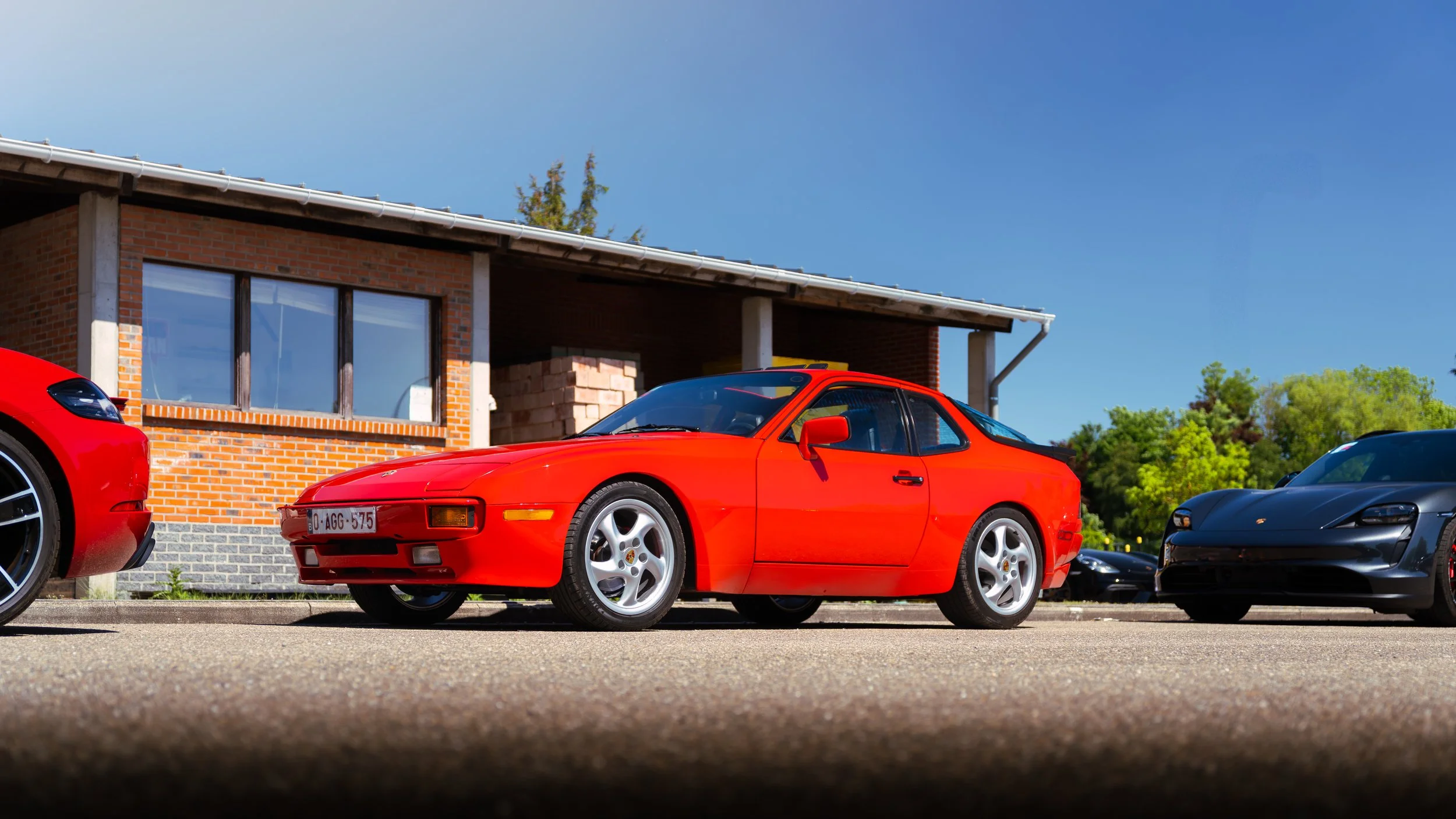 A red sports car parked on a driveway with other cars near a brick house under a clear blue sky.
