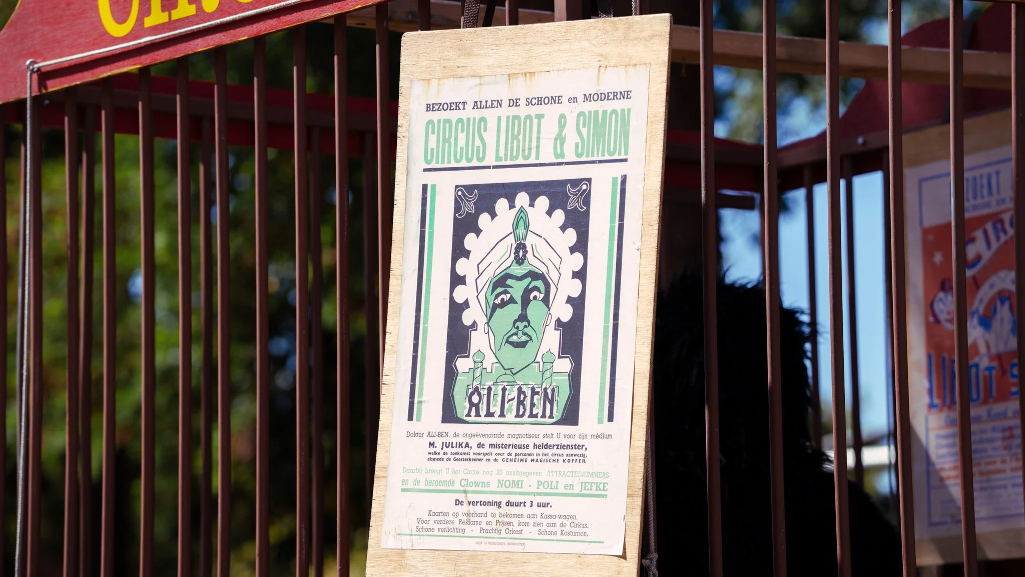 A vintage-style circus poster advertising Circus Libot & Simon with a stylized illustration of a face in green and black, hanging on a wooden board among metal bars.