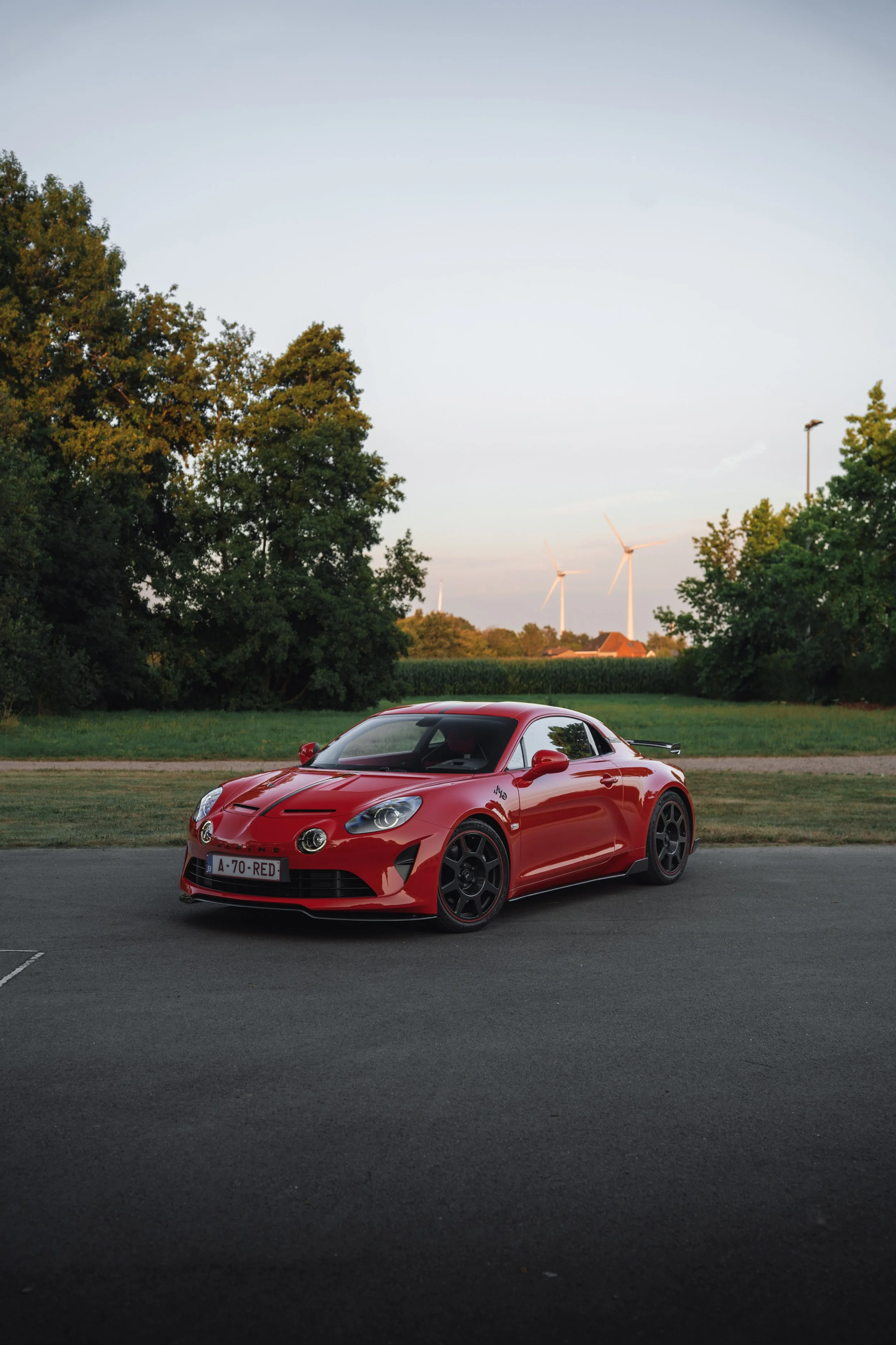 A red sports car parked on a paved lot with grassy and tree-lined background, wind turbines visible in the distance, and a clear sky.