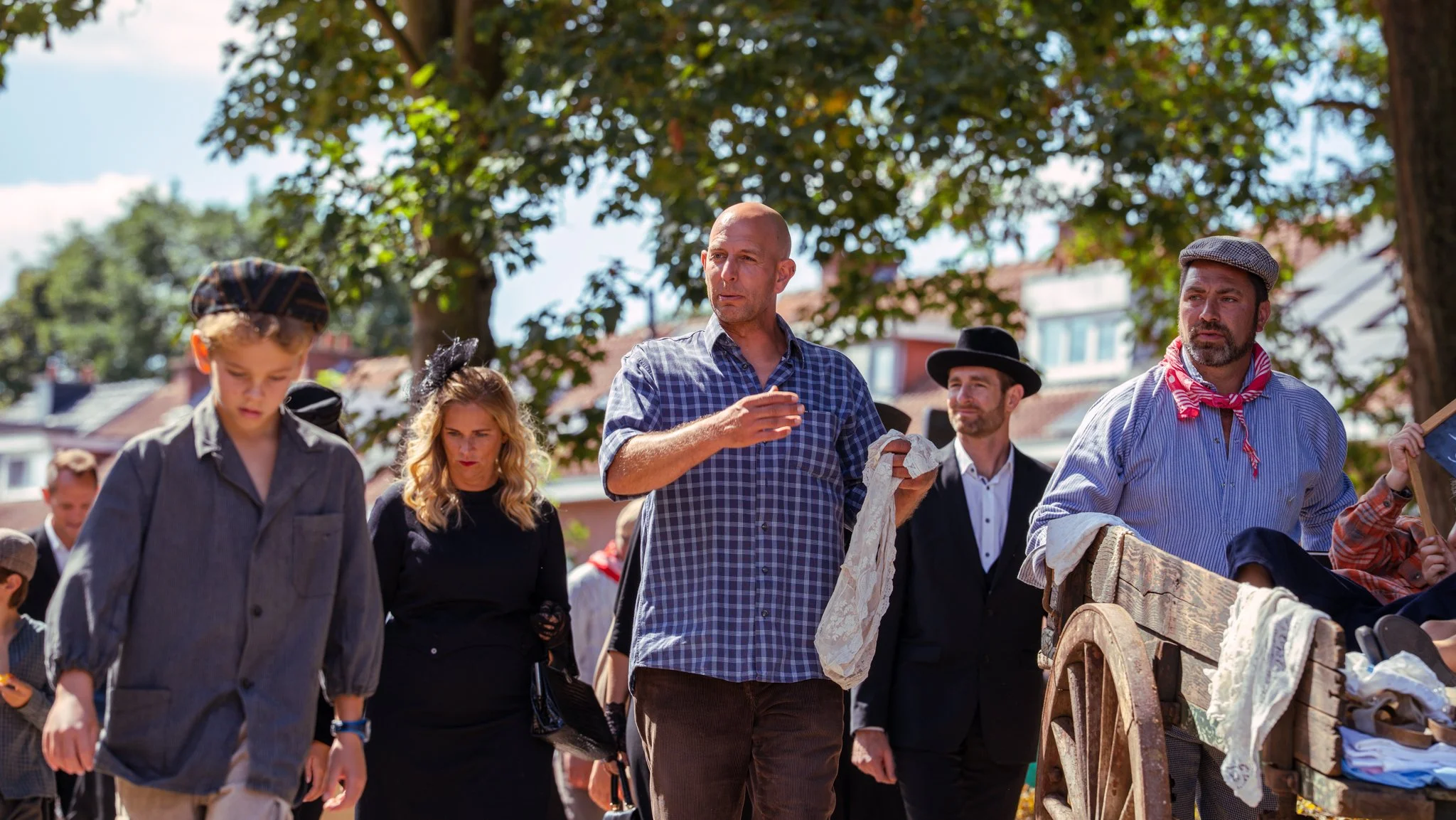 Group of people dressed in vintage clothing at an outdoor market or fair, with trees and buildings in the background.