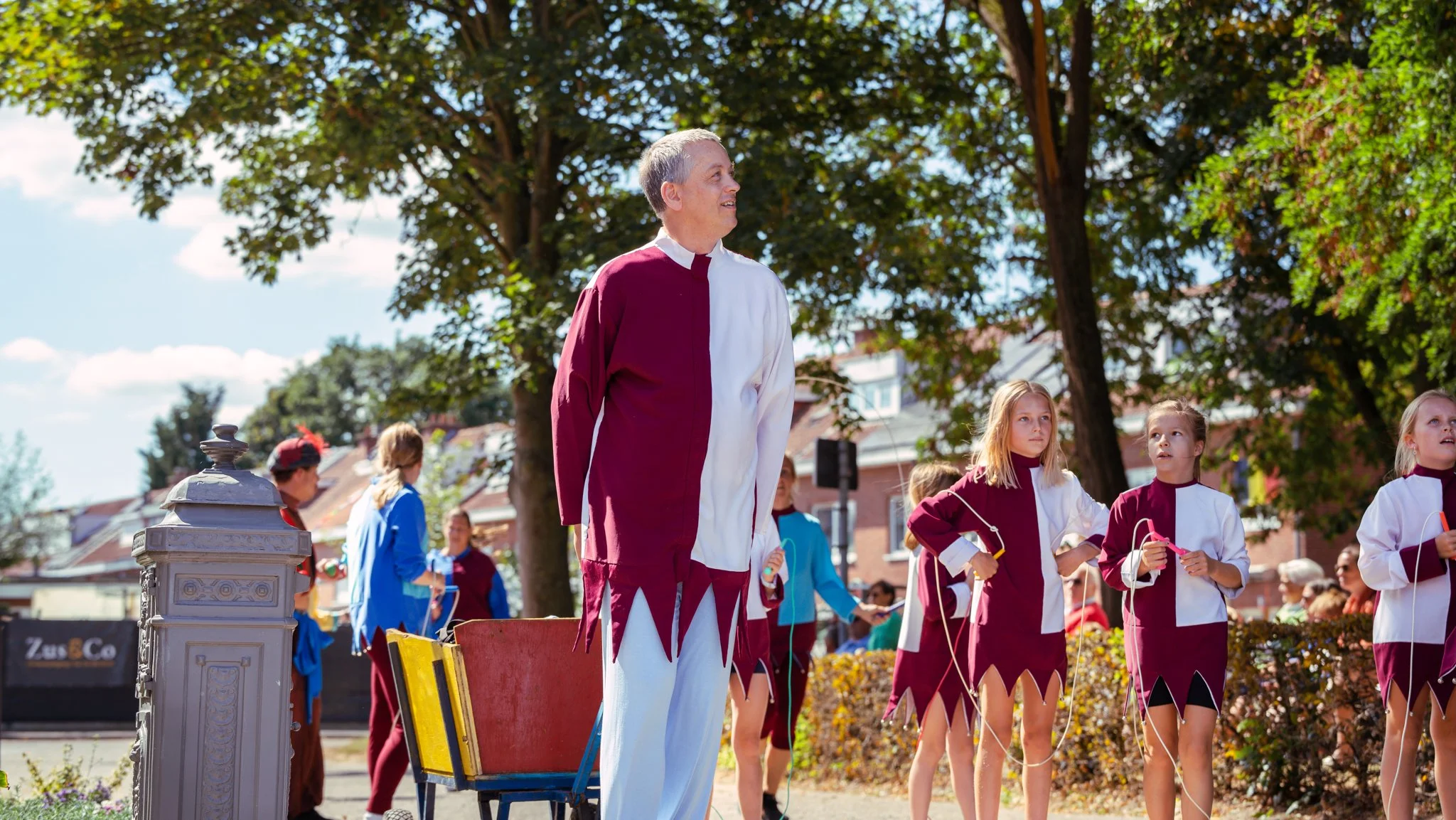 A man dressed as a jester standing among children in medieval-style costumes during an outdoor event on a sunny day with trees and buildings in the background.