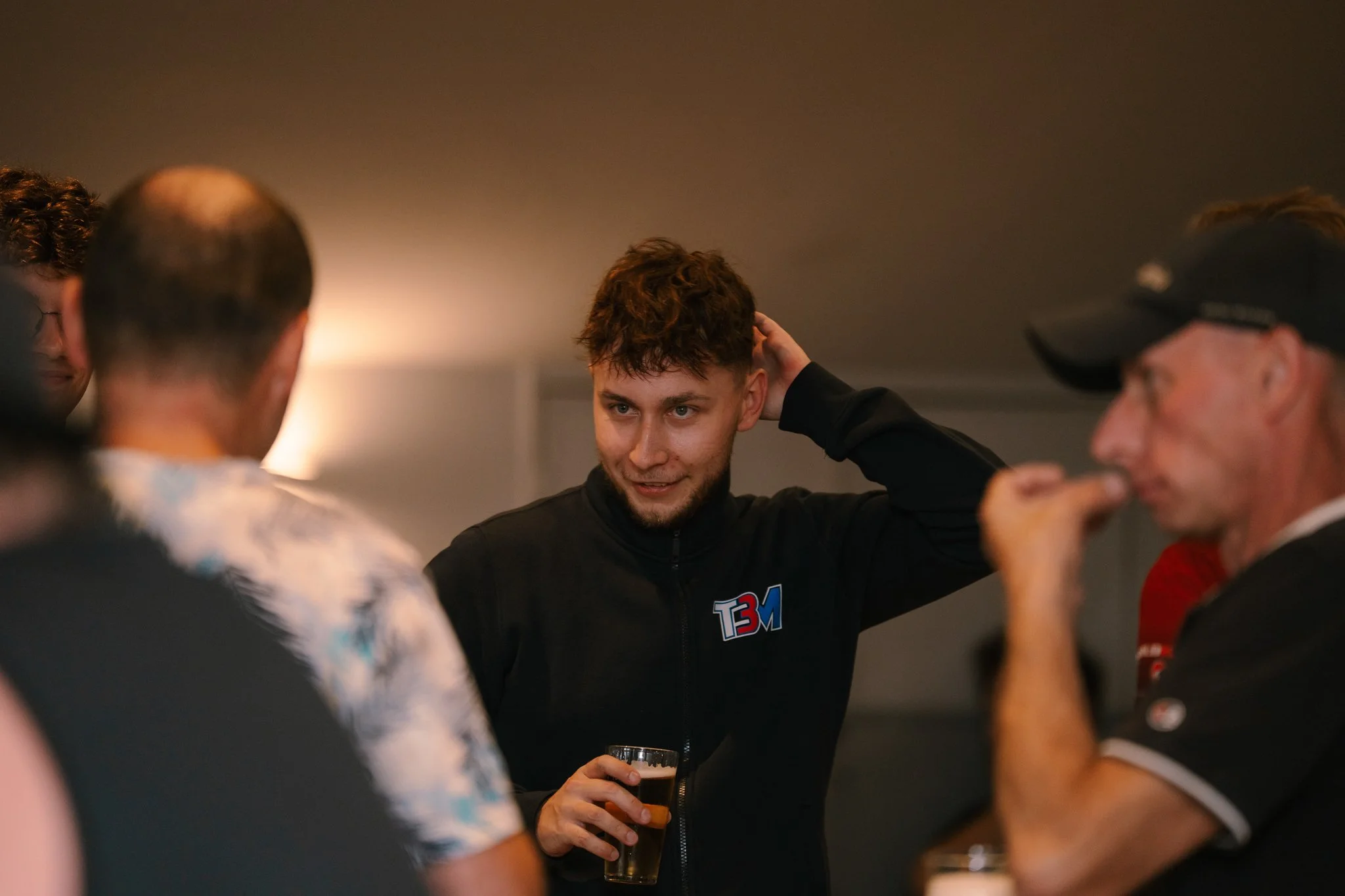 Young man with curly hair and a beard holding a beer, smiling, in conversation with others at a social gathering indoors.