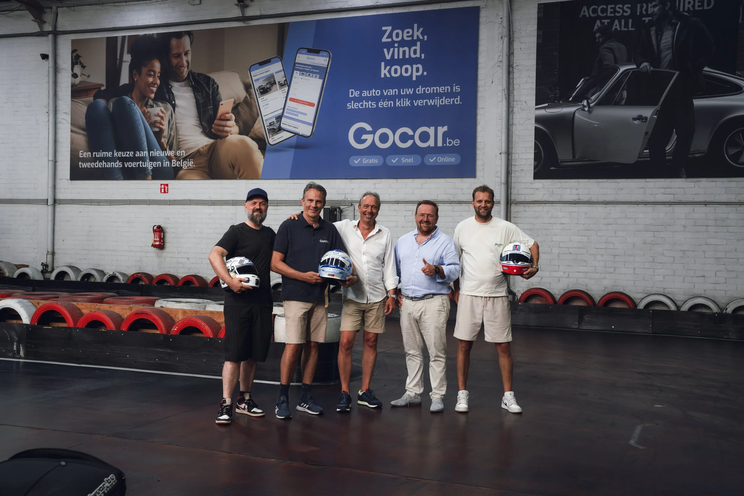 Five men are posing together in a racing facility holding helmets in front of a large wall with advertisements. The wall features a large poster with a couple looking at a phone, promoting a car buying app, and an image of a man with a silver car. Th