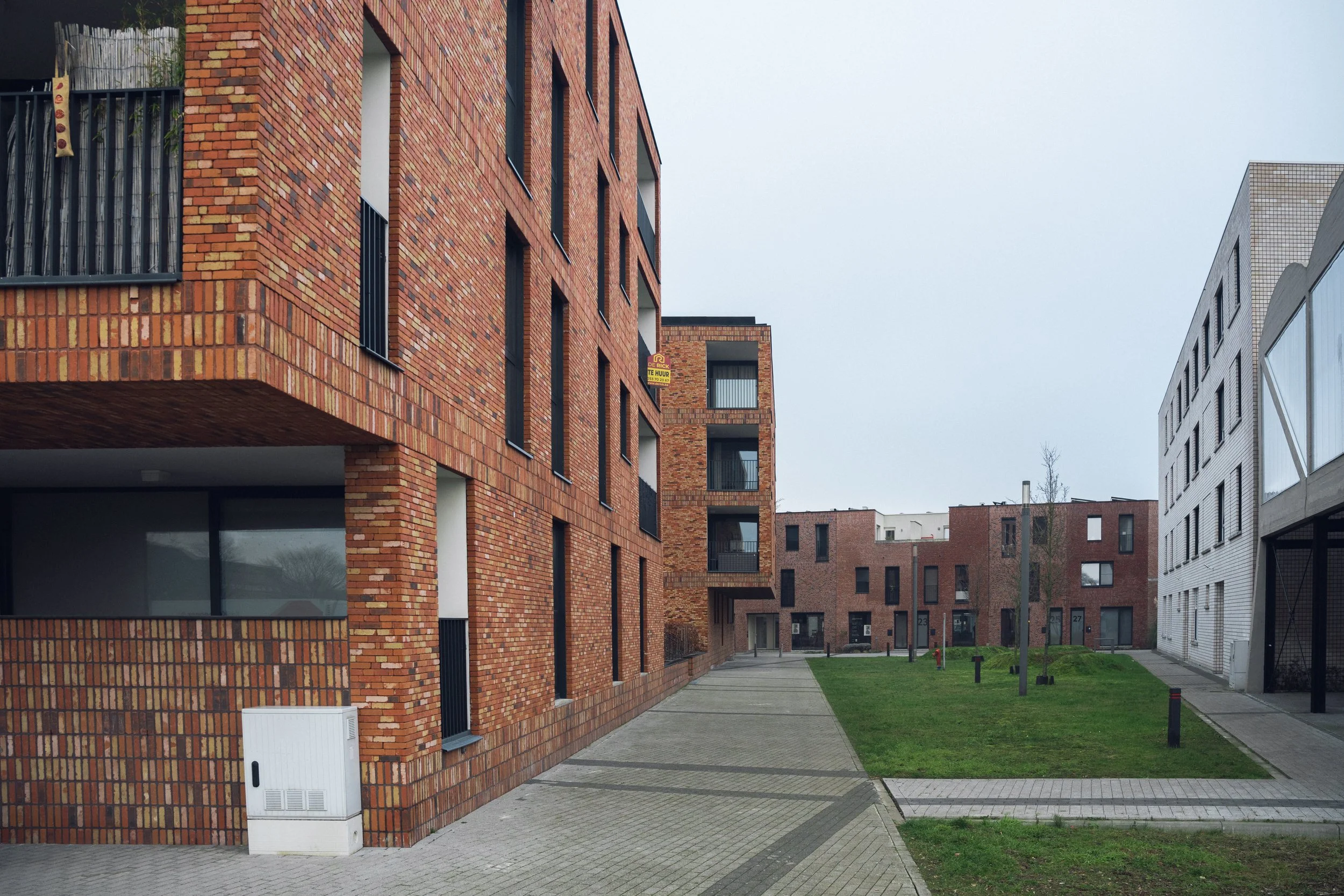 A modern residential complex with red brick buildings, some with balconies, and a small green courtyard with trees and a paved pathway.