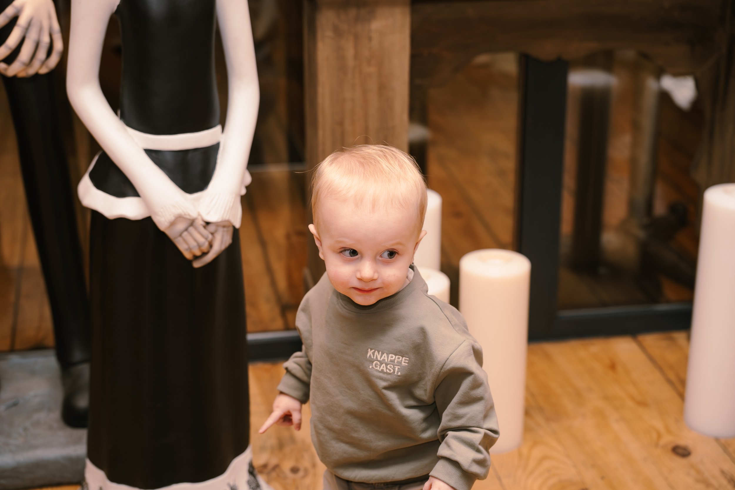 Young child with reddish blonde hair and gray shirt looking to the side, standing near display mannequins in a wooden-floored room.