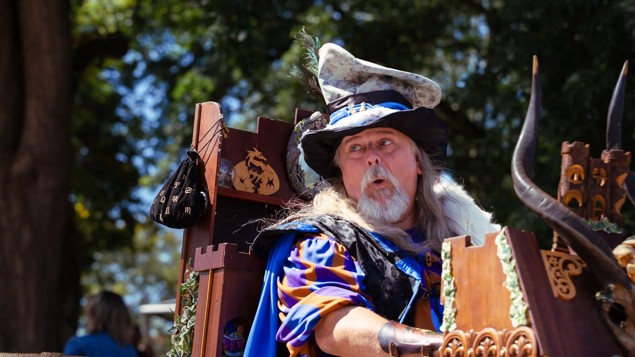 A man dressed as a wizard with a long white beard, wearing a large, elaborate hat, sits on a wooden prop with fantasy-themed decorations, outdoors with trees in the background.
